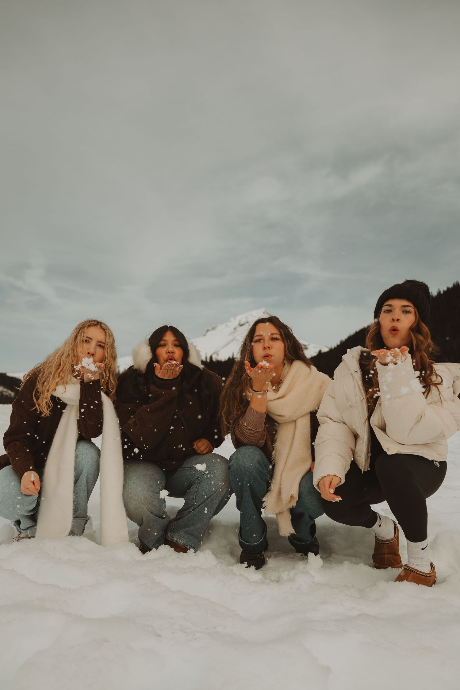 Four women crouch in snow blowing snowflakes toward camera with snowy mountain backdrop in winter.
