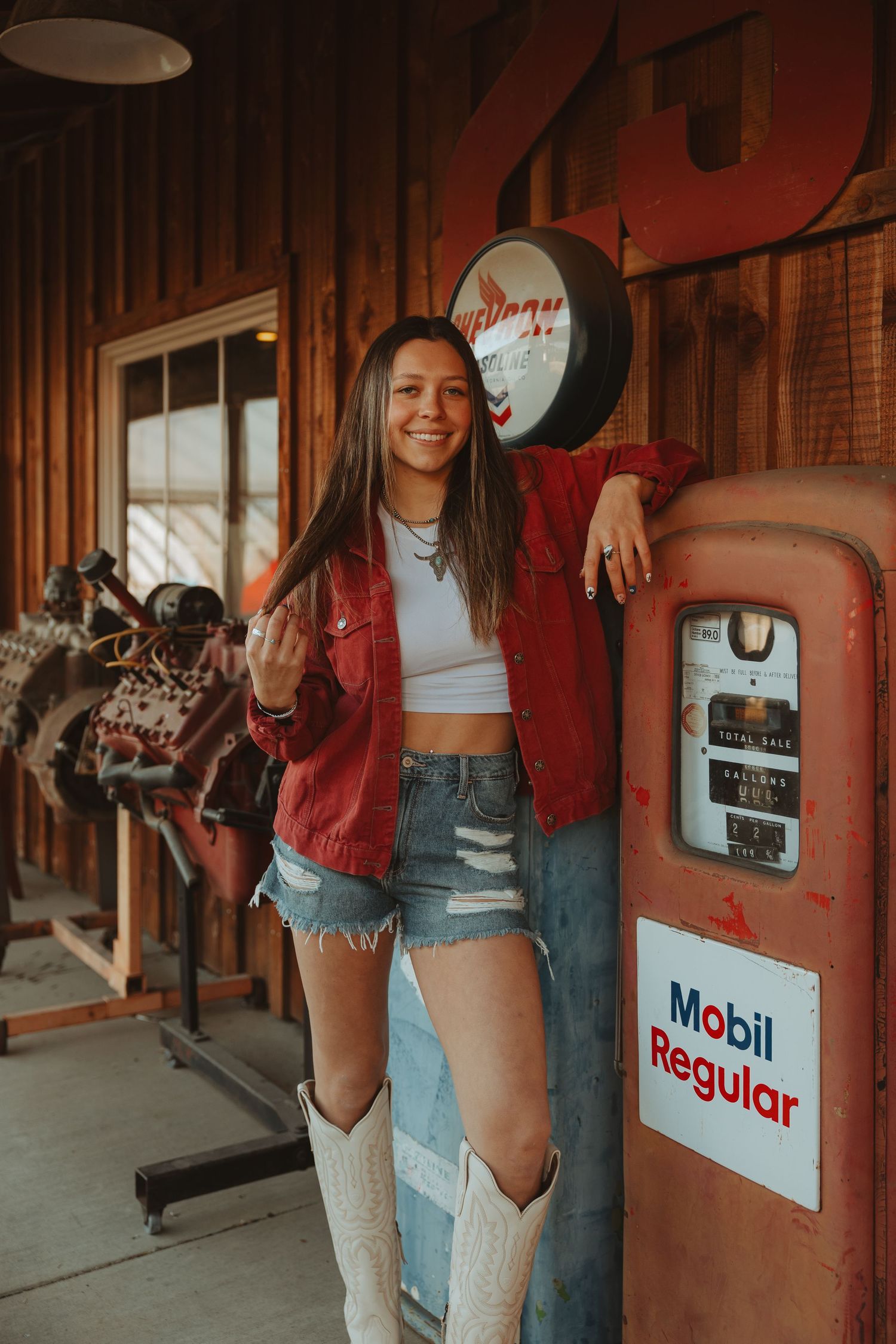 Young woman in red jacket, denim shorts, and white cowboy boots leaning on a vintage Mobil Regular gas pump.
