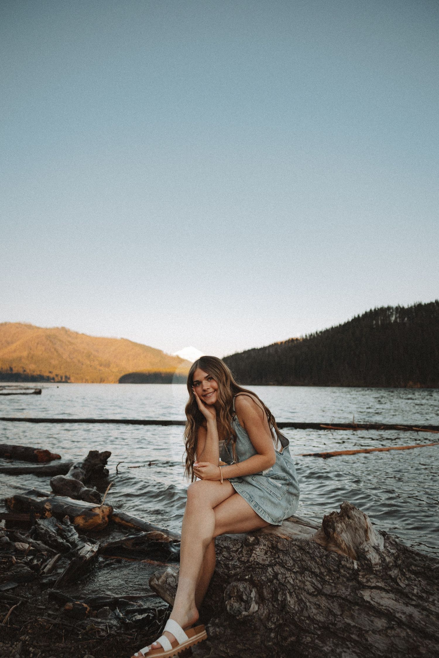 Young woman in denim dress sits on driftwood by a mountain lake at sunset, smiling warmly at camera.