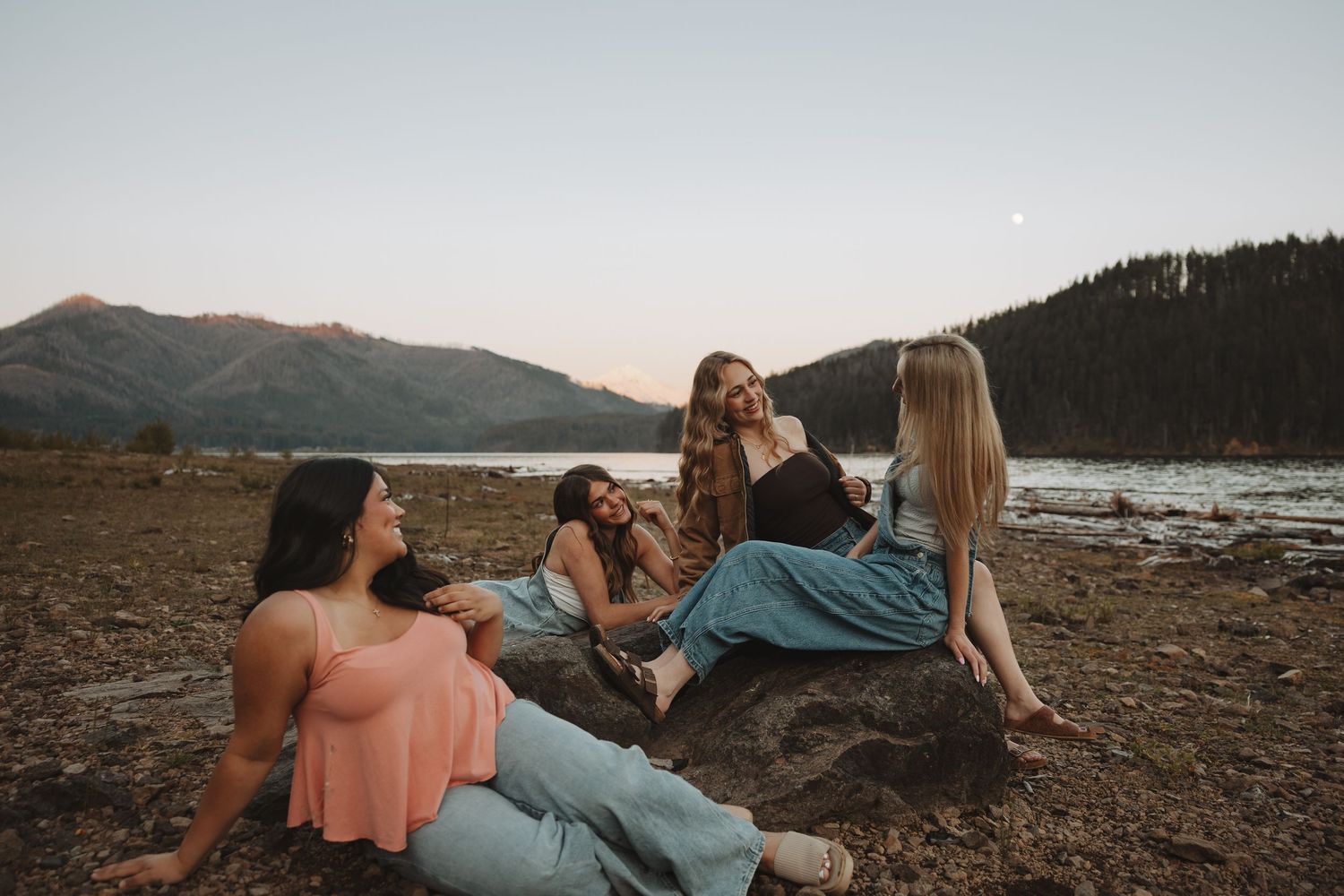 Four young women relaxing on rocks by mountain lake at dusk with crescent moon in sky.