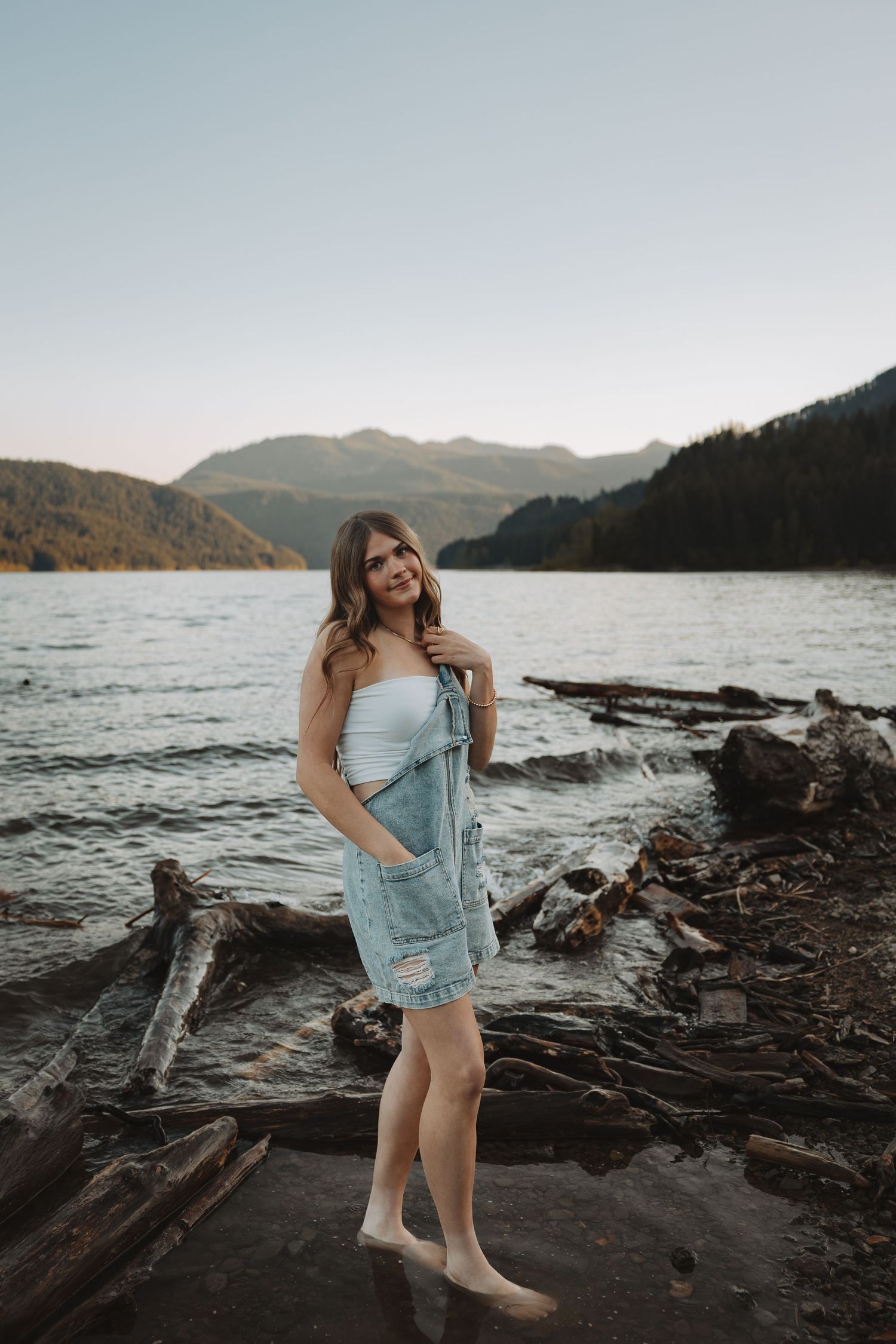 Young woman in white top and denim jacket standing on driftwood by scenic mountain lake at dusk.