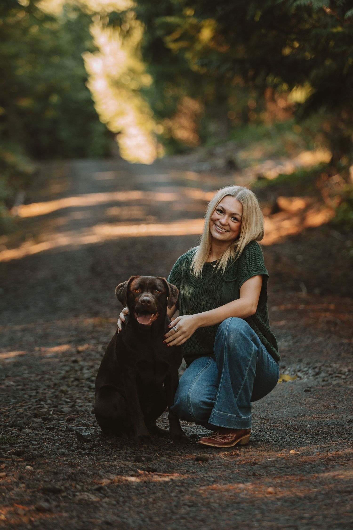 A chocolate Labrador and its companion share a moment on a sunlit forest path during golden hour.