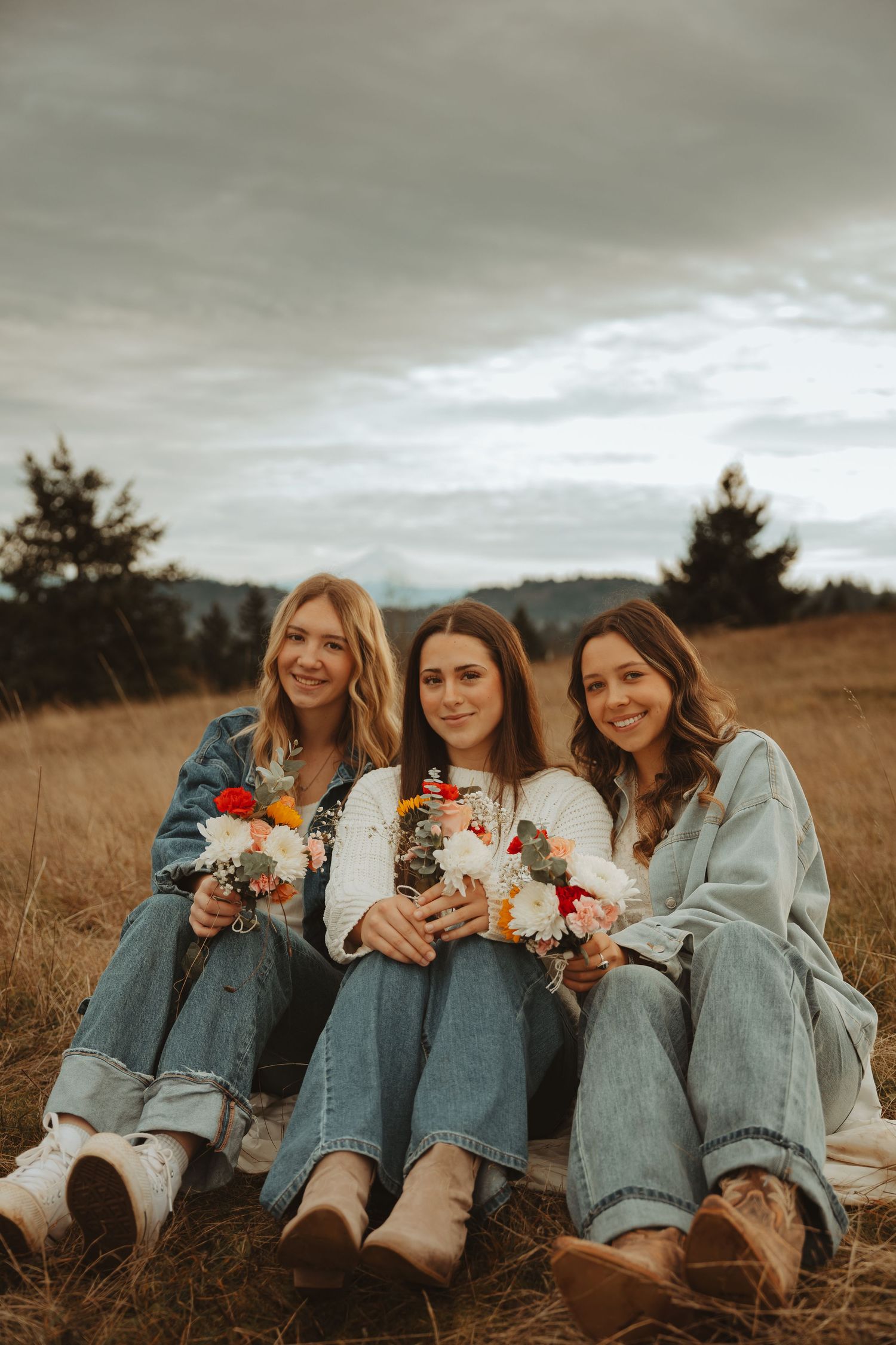 Friends wearing coordinated denim and white outfits pose with flowers in a rustic field setting.