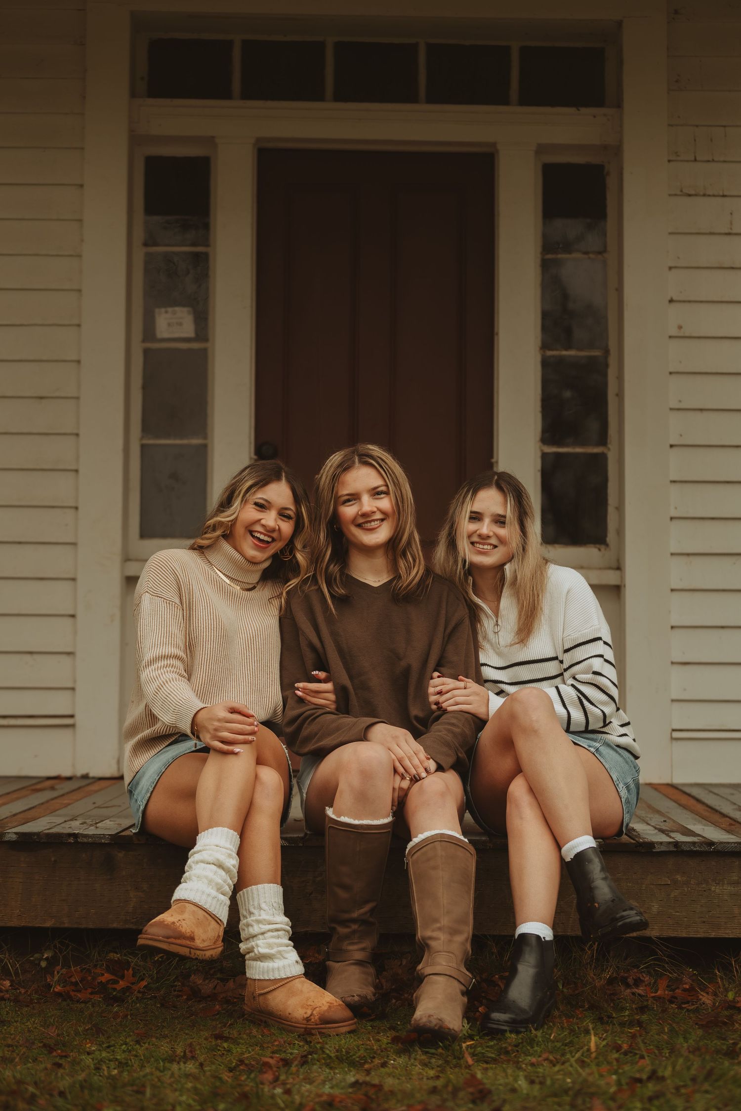 Three friends sitting casually on wooden porch steps wearing cozy sweaters and boots on an autumn day.