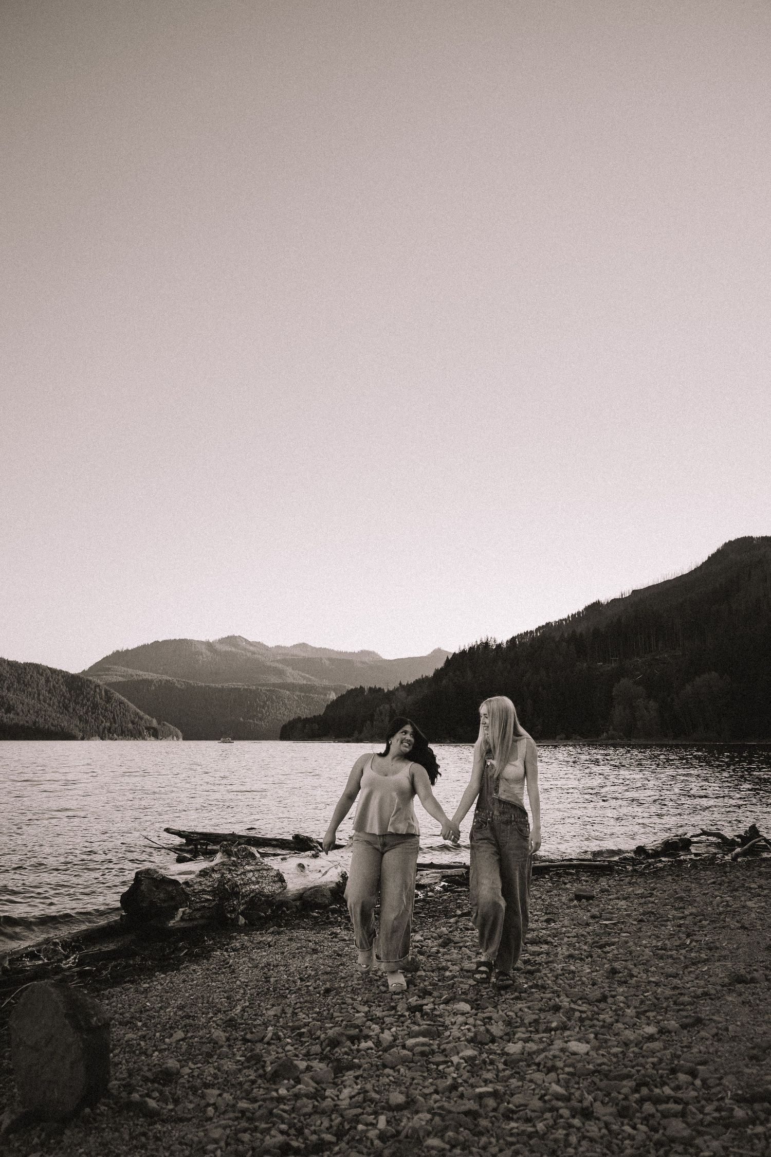 Black and white photo of two women holding hands walking along a rocky mountain lakeshore.
