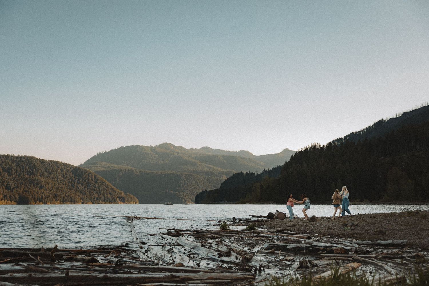 Group of friends walking along a rocky lakeside beach with mountain and forest backdrop at dusk.