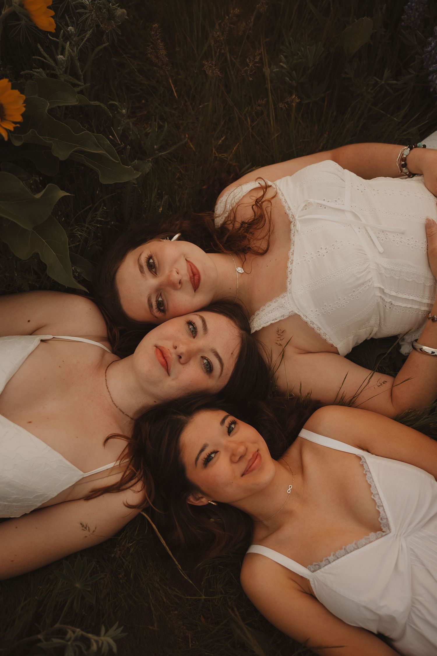 Three young women in white dresses lying together in a lush garden with sunflowers, gazing at the camera.