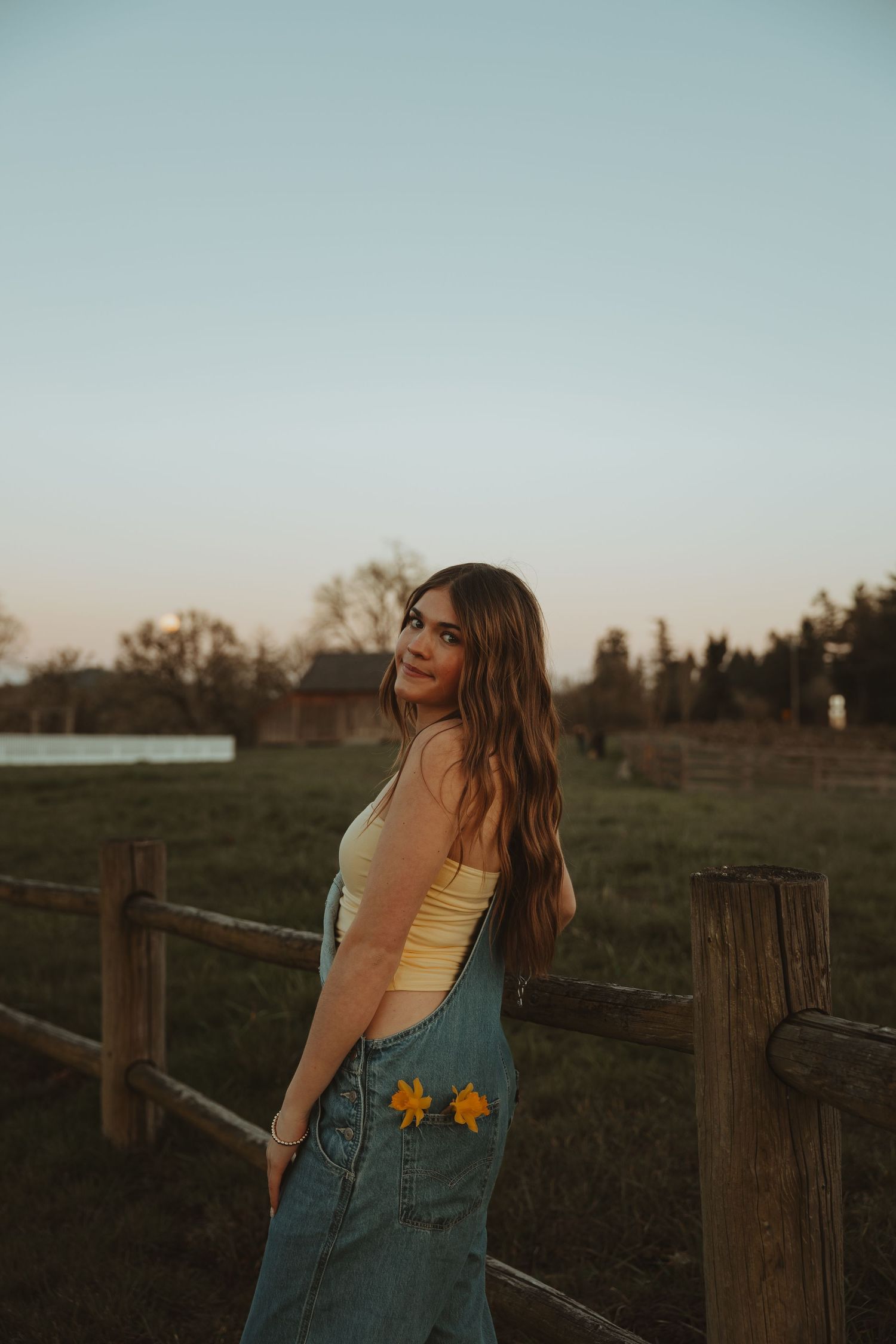 Young woman in denim overalls and yellow top poses by rustic wooden fence at golden hour sunset.