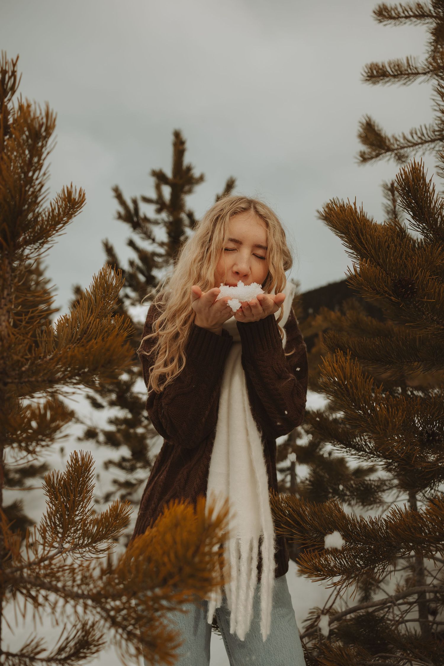 Blonde woman blowing snow from hands surrounded by pine trees in winter.