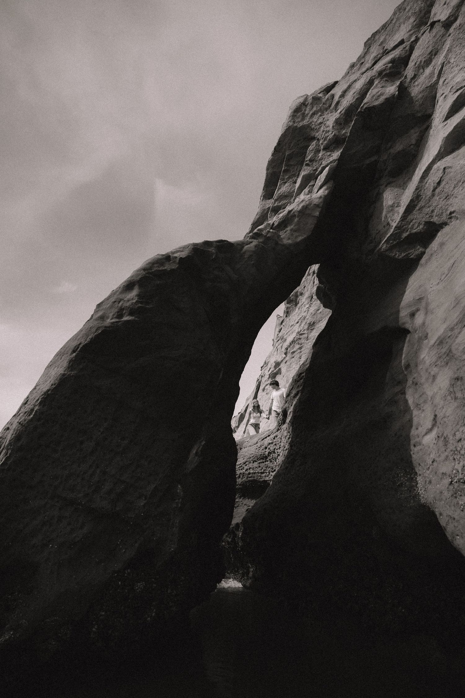 Black and white photo of dramatic sandstone rock arch with cloudy sky above.