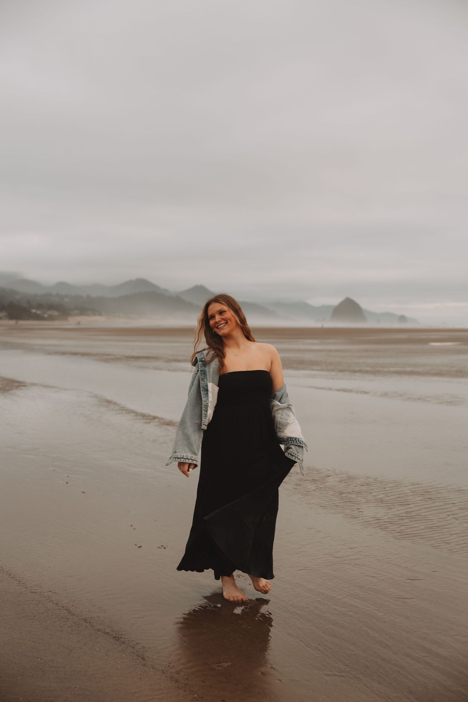 A moody scene on a misty beach with a figure in black dress and denim jacket against an overcast sky.