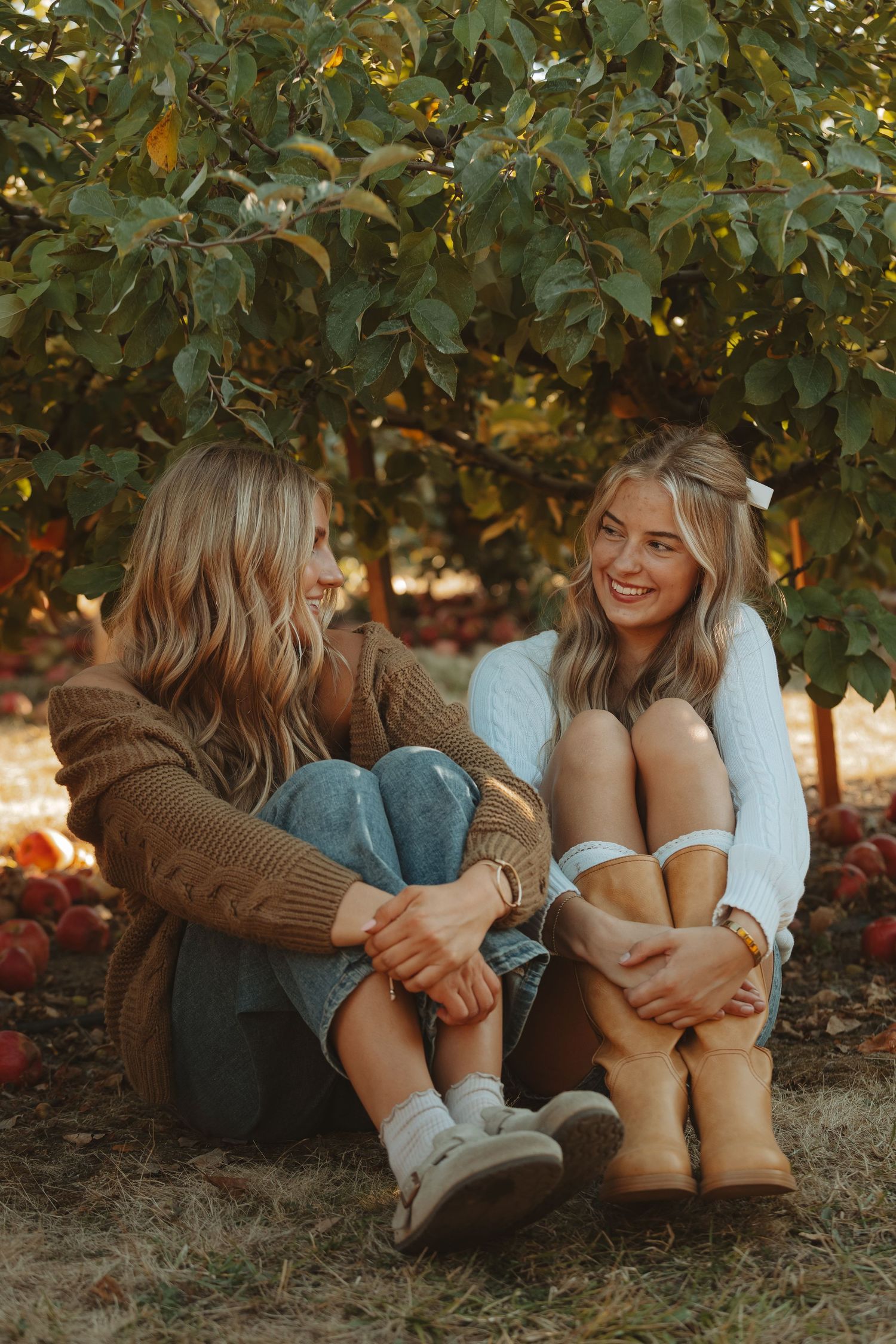 Two friends share joyful moments while sitting together on grass under autumn trees in casual outfits.