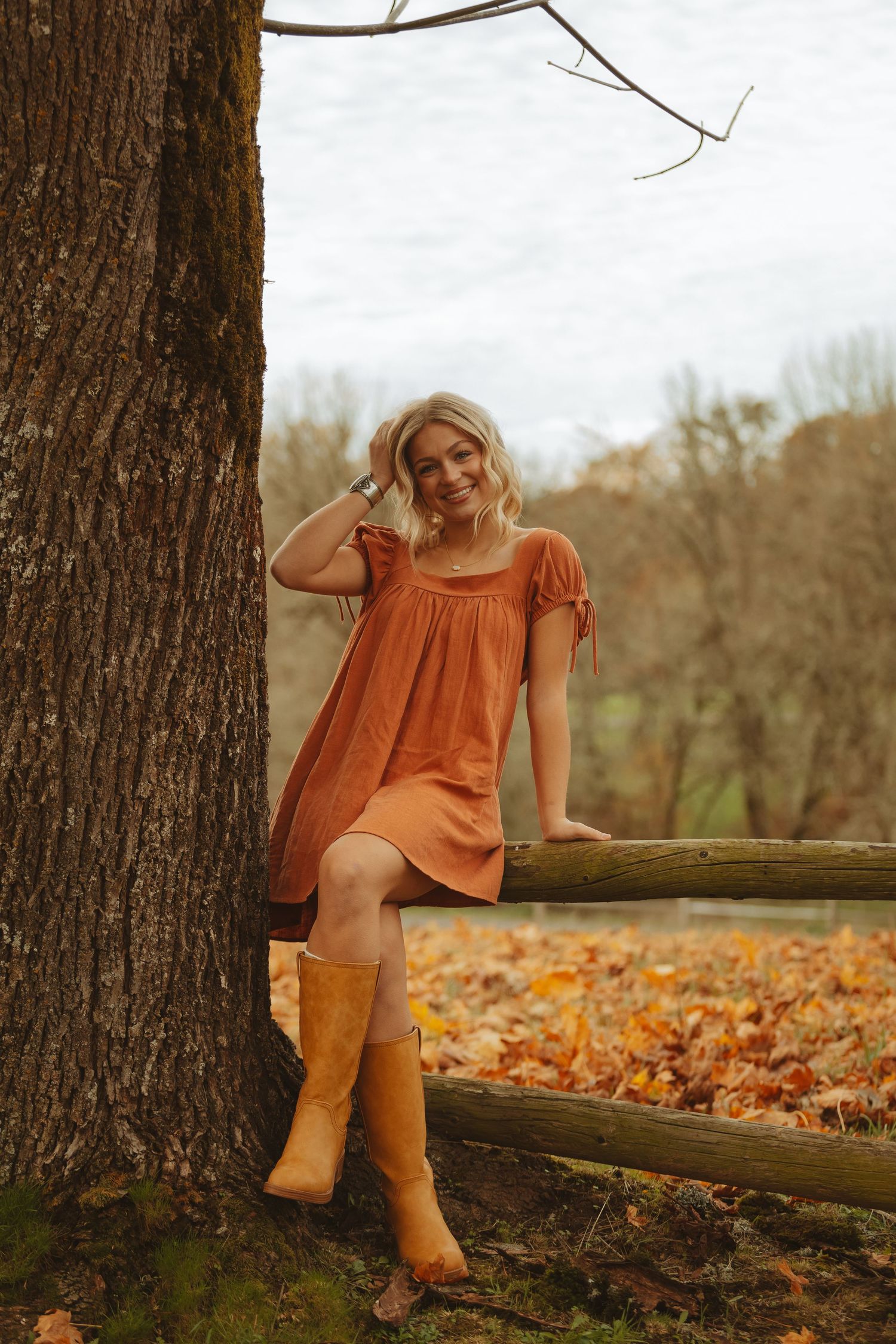 Woman in orange dress and boots poses by fallen log surrounded by autumn leaves.