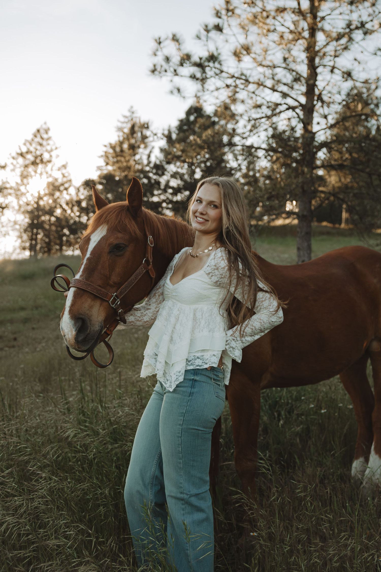 Young woman in white lace top and jeans smiling beside a chestnut horse in a sunlit green field.