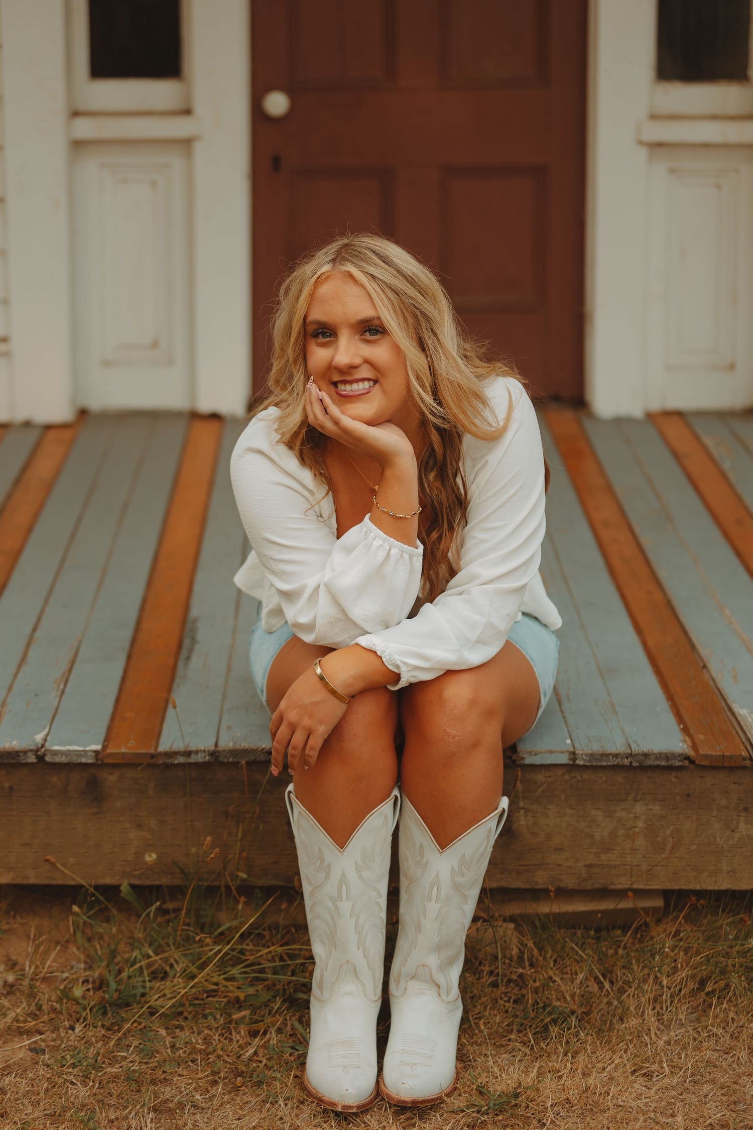 Smiling blonde woman in white top and cowboy boots sitting on vintage porch steps of old building.