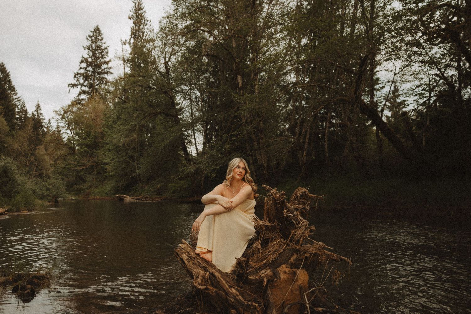 Pregnant woman in cream dress sitting on driftwood by a river surrounded by lush green forest trees.