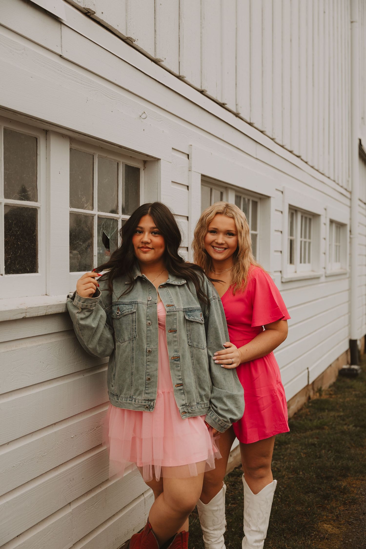 Two friends in denim jacket and pink dress pose together against white building exterior.