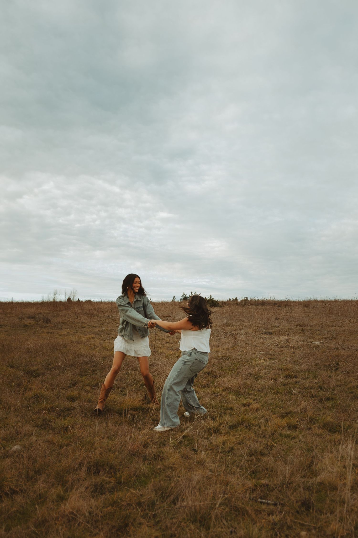 Two friends in white sweaters playfully dance together on a brown grassy hillside under cloudy skies.