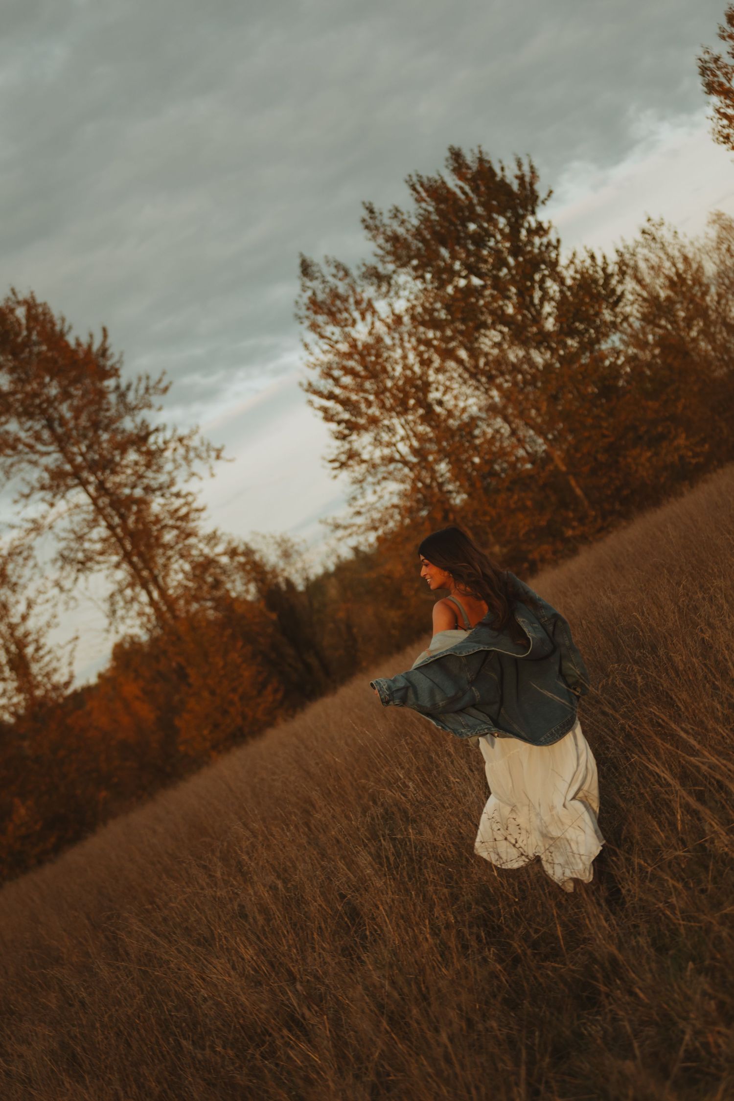 Person spinning in white dress and denim jacket among tall grass at dusk.