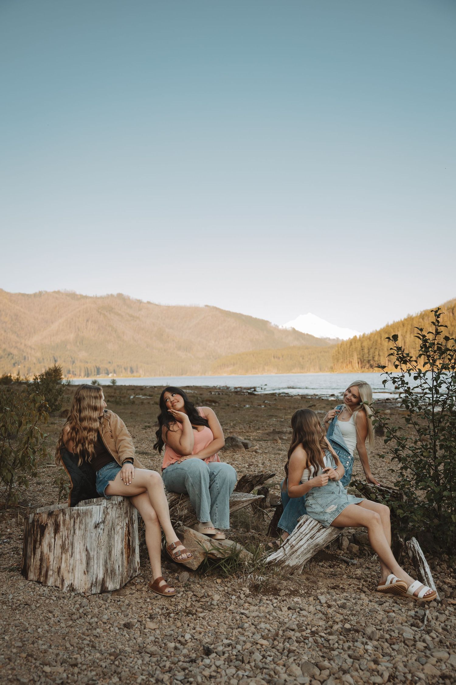 Four women sitting on logs near a mountain lake at golden hour in a scenic outdoor setting.