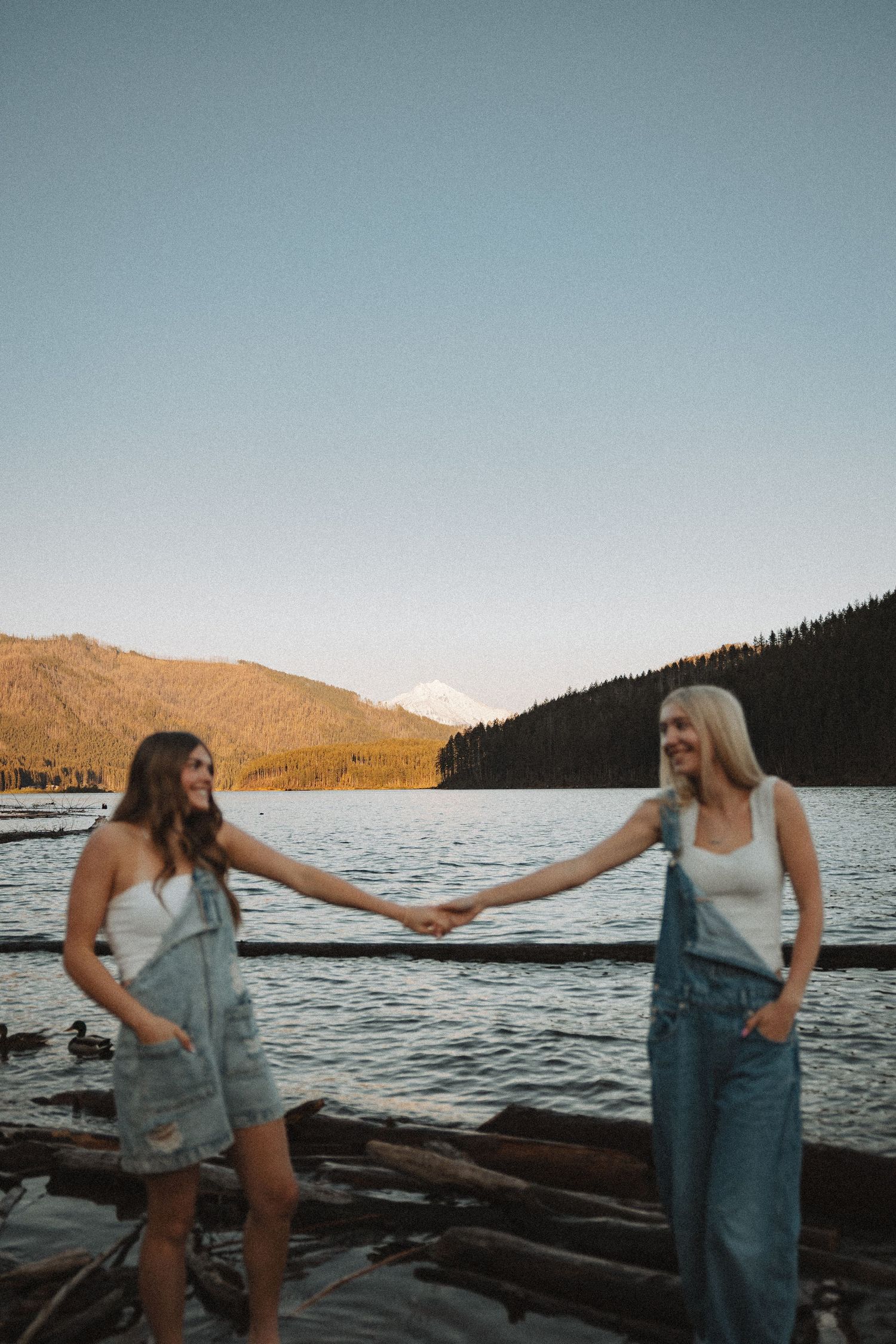 Two young women holding hands on a rocky lakeside with a snow-capped mountain visible across the lake.