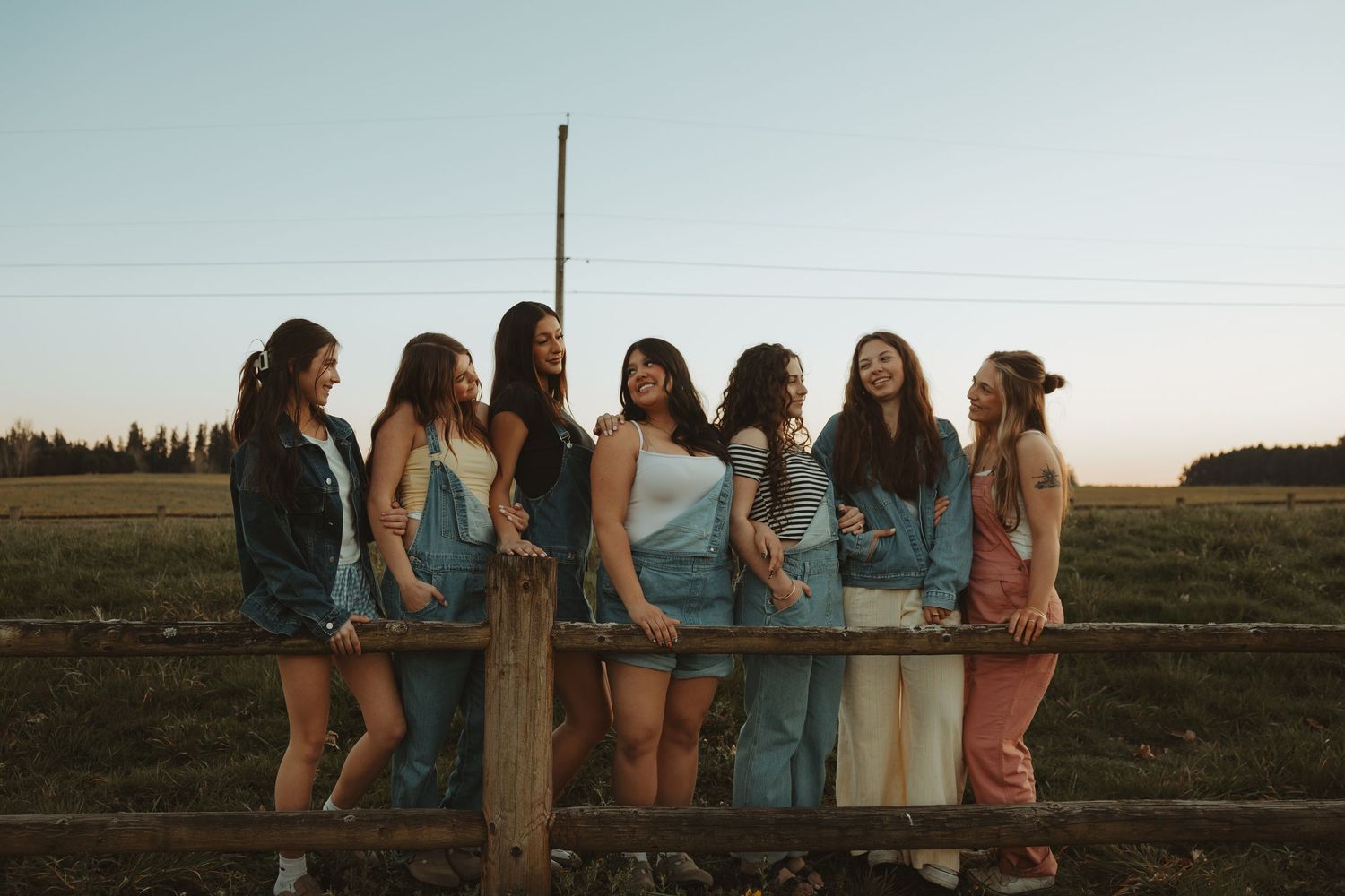 Seven young women leaning on a rustic wooden fence at sunset in a scenic outdoor field setting.