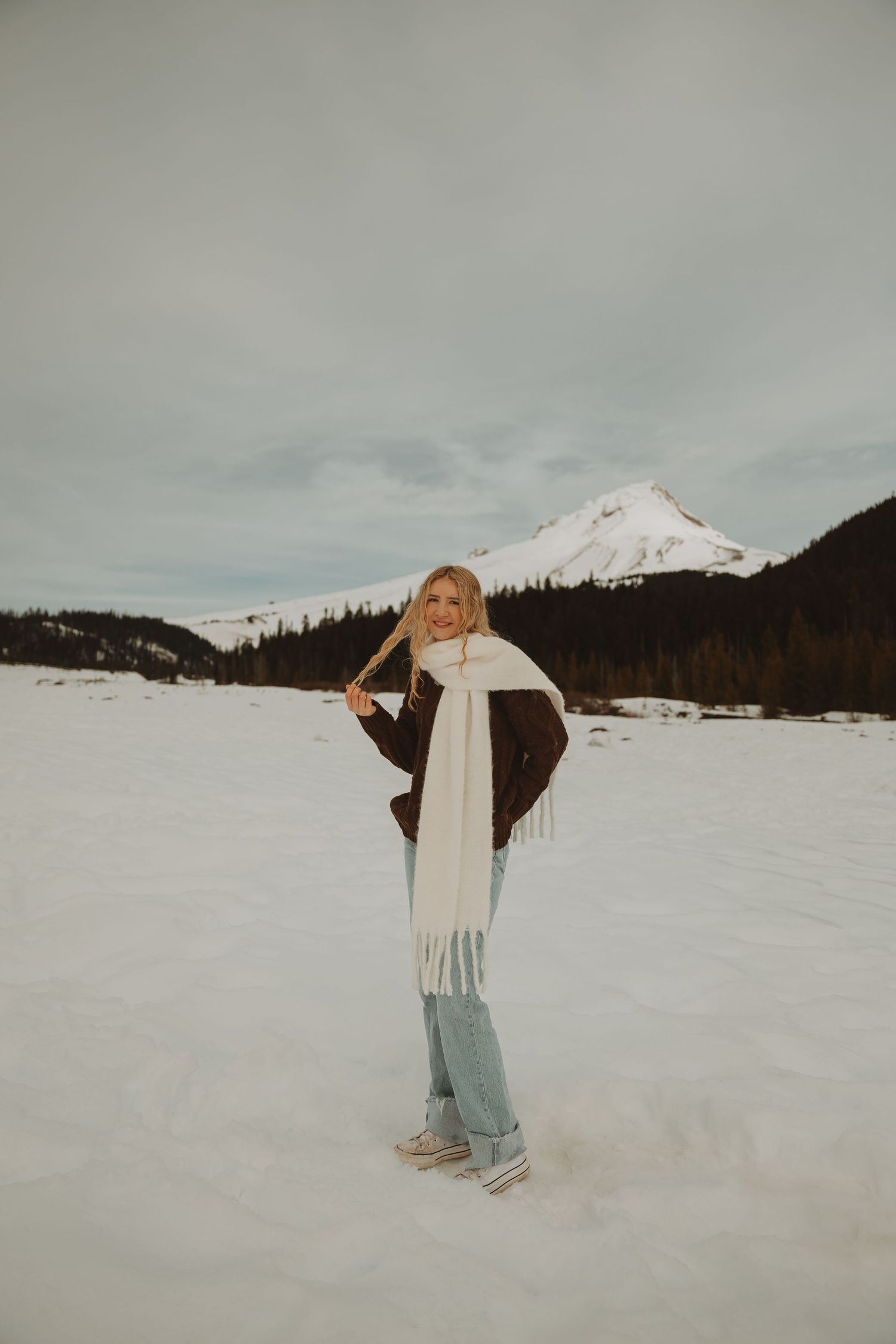 Blonde woman in brown jacket and white fringe scarf poses on snowy field with mountain behind her.