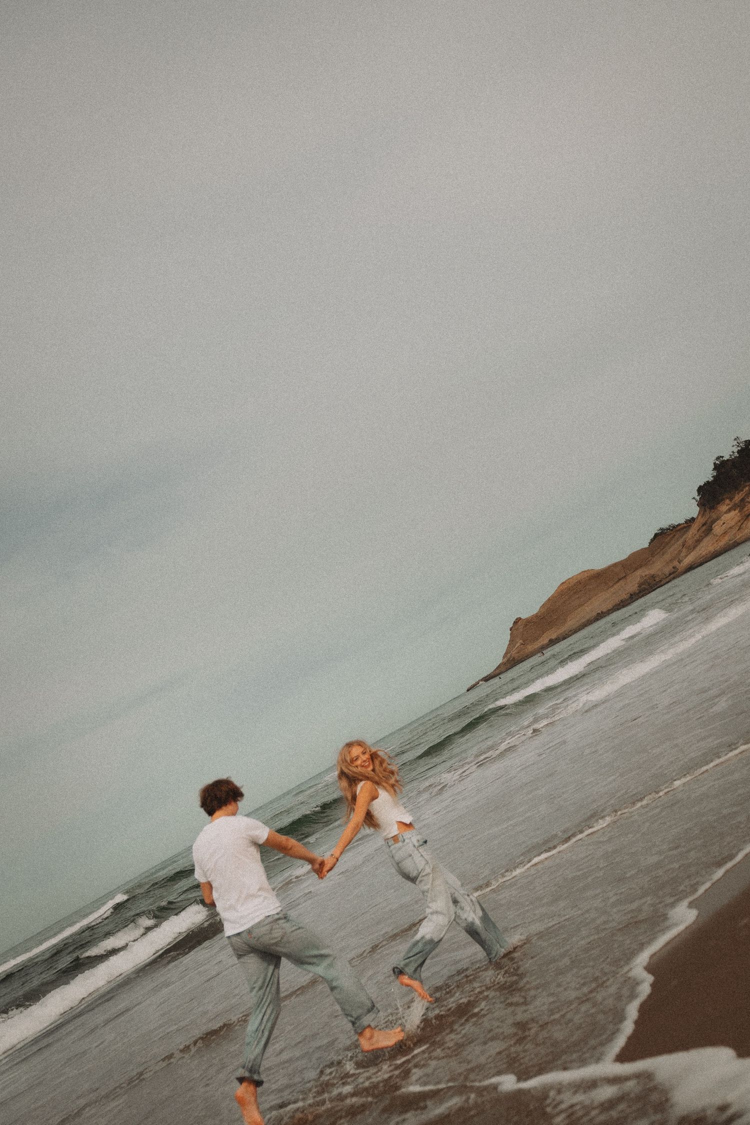 Couple holding hands and dancing barefoot along the shoreline with coastal cliffs visible in the background.