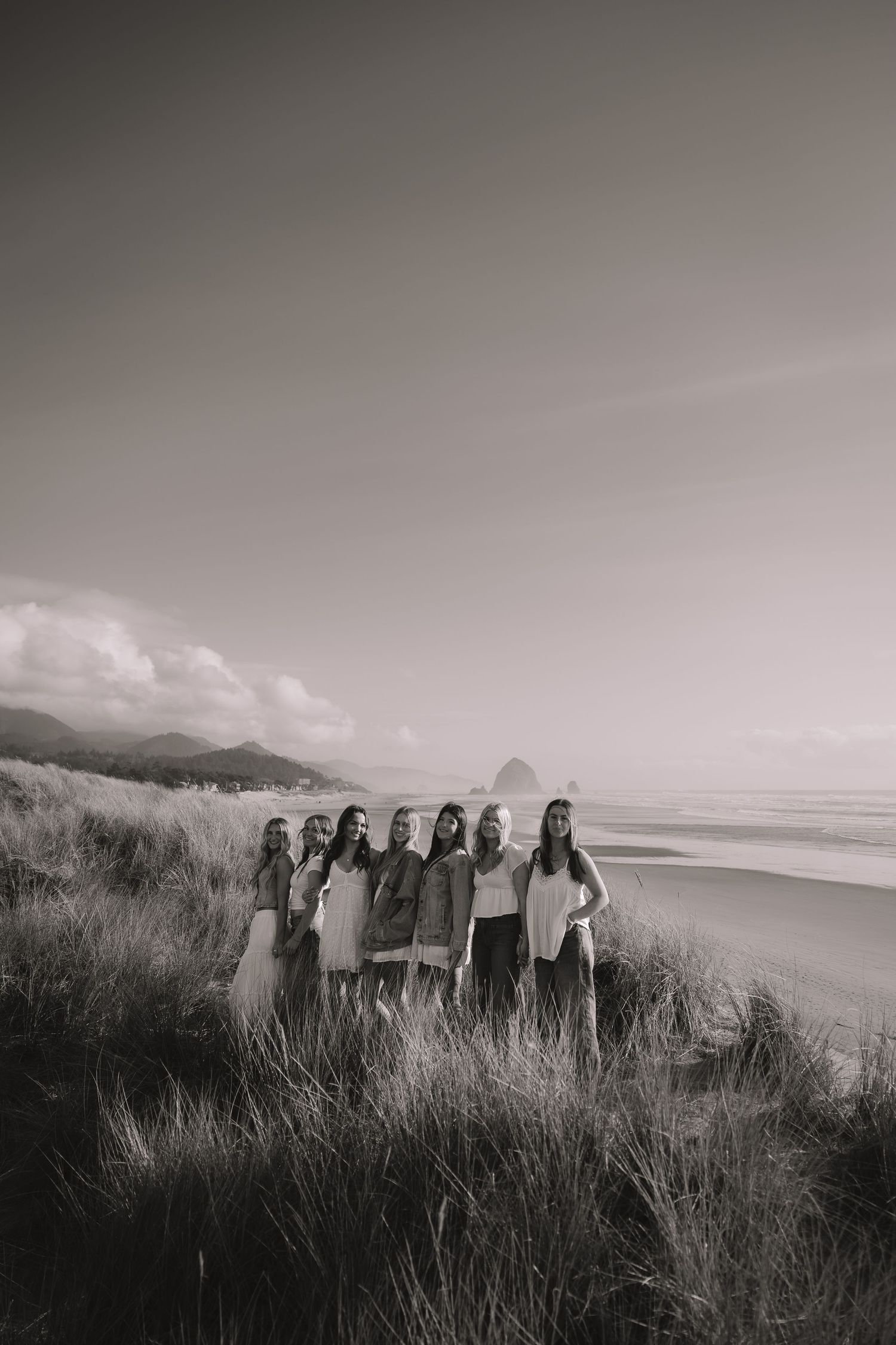 Black and white photo of seven women standing in beach dunes with Haystack Rock backdrop.