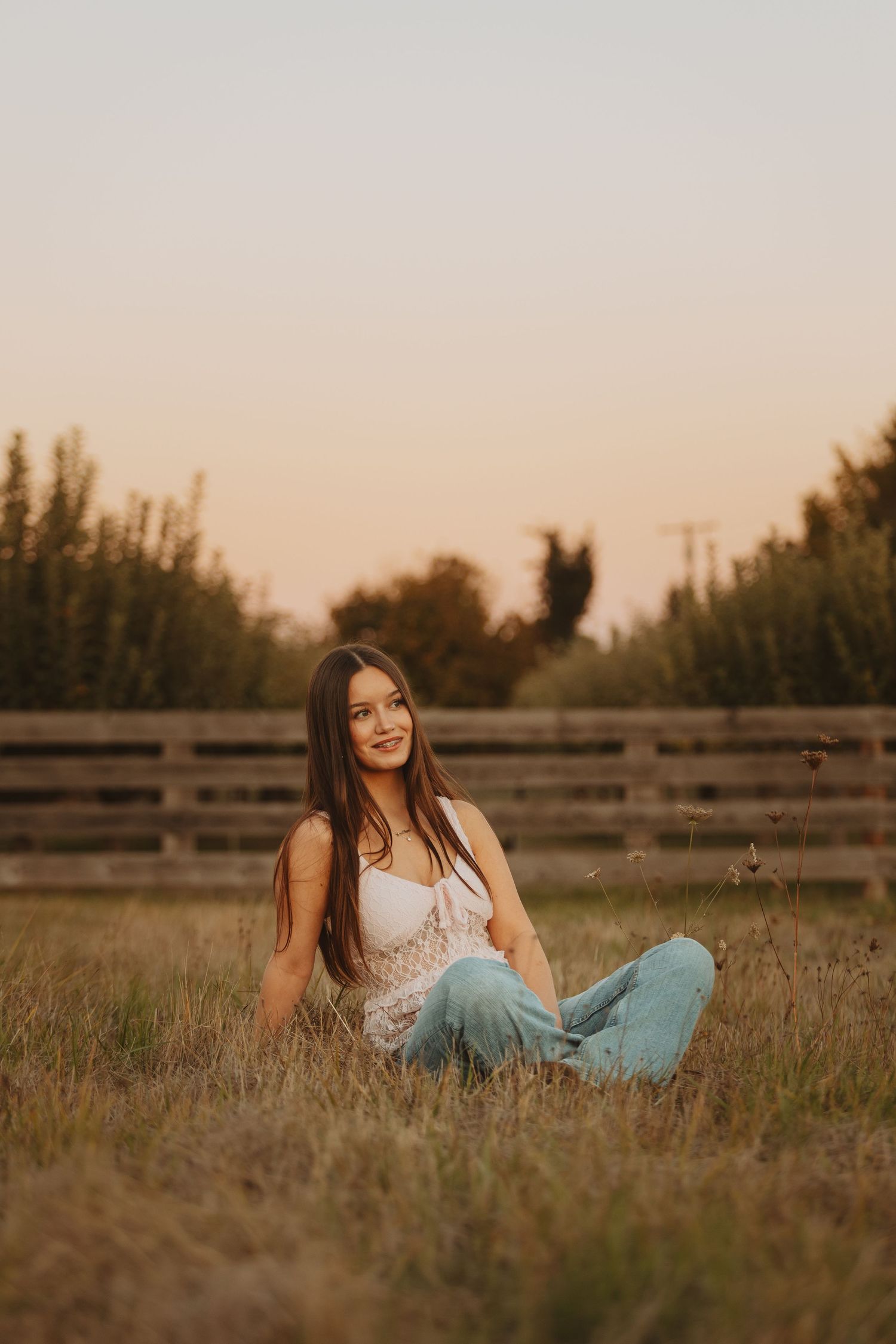 Young woman sitting cross-legged in a golden field at sunset, wearing a white lace top and blue jeans near a wooden fence.
