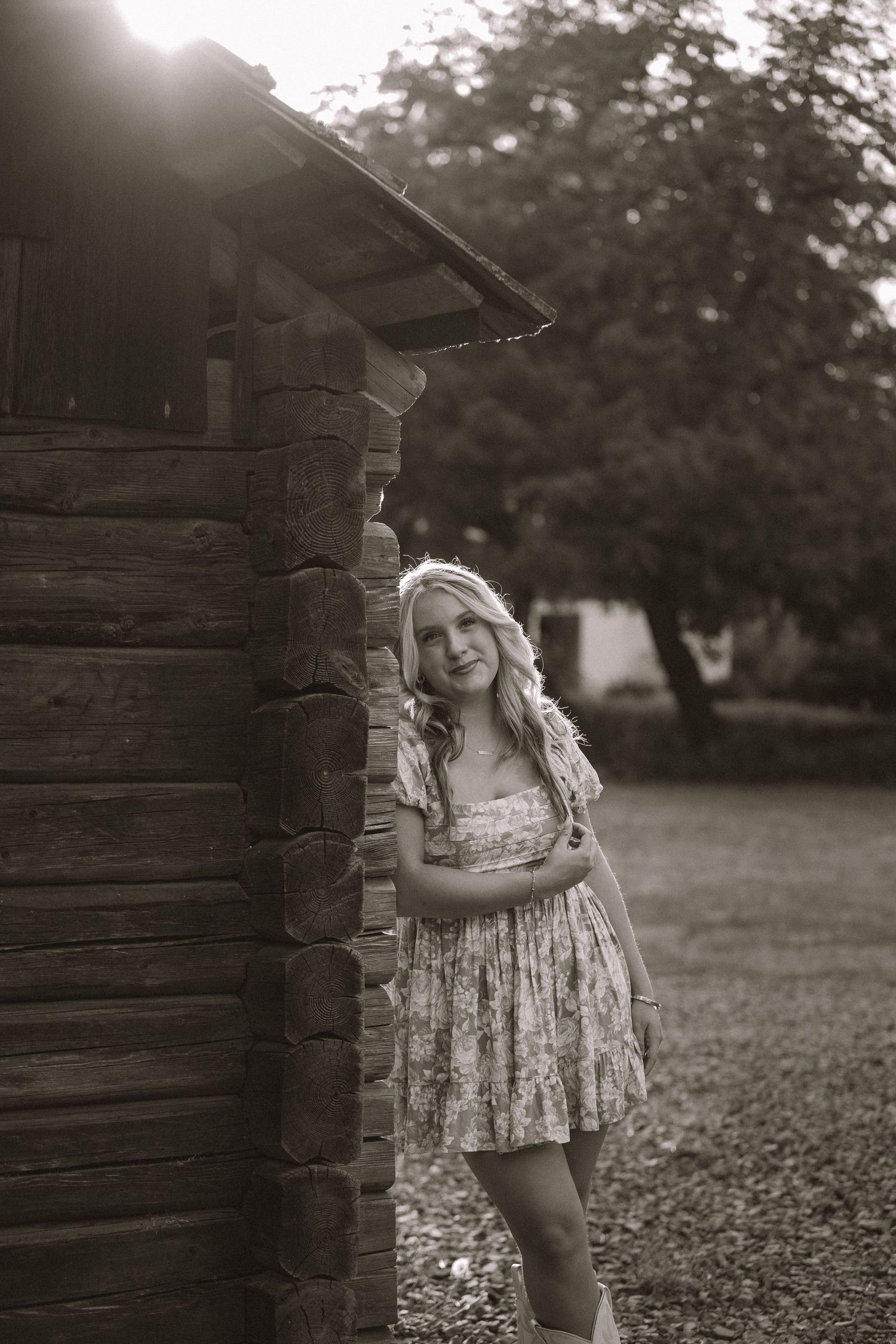 Smiling blonde teen girl in floral dress leaning against rustic log cabin corner in sepia toned outdoor portrait.