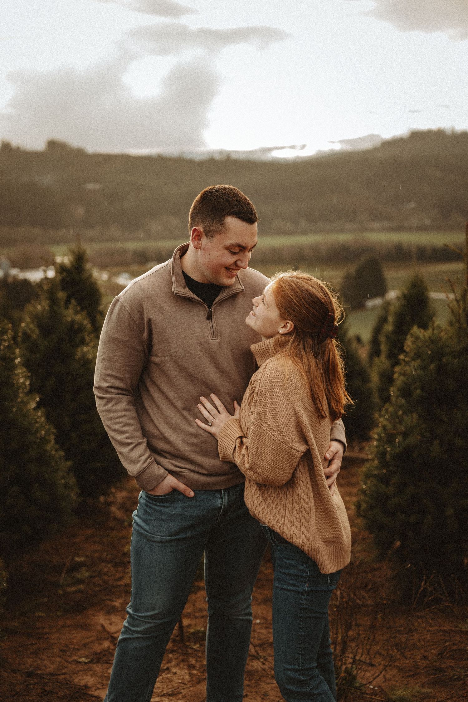 A couple in matching beige sweaters and jeans share an intimate moment in a tree farm at sunset.