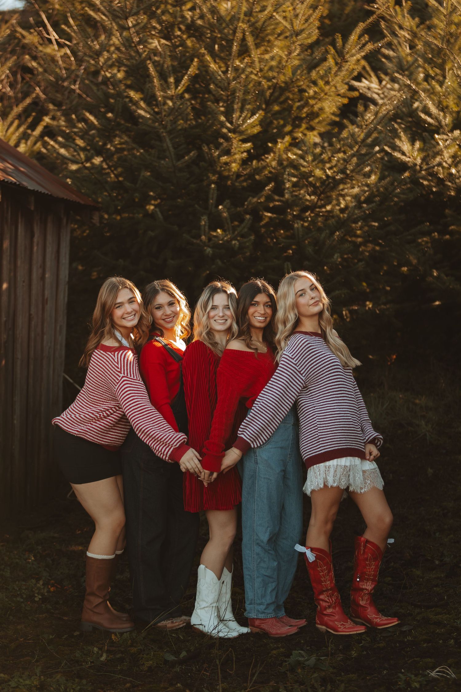 Friends in cozy winter sweaters pose together by wooden barn at sunset.