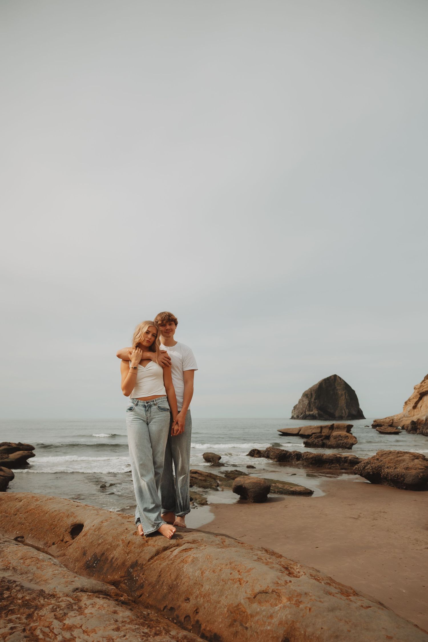Romantic couple embracing on rocky beach with haystack rock and ocean waves in background.