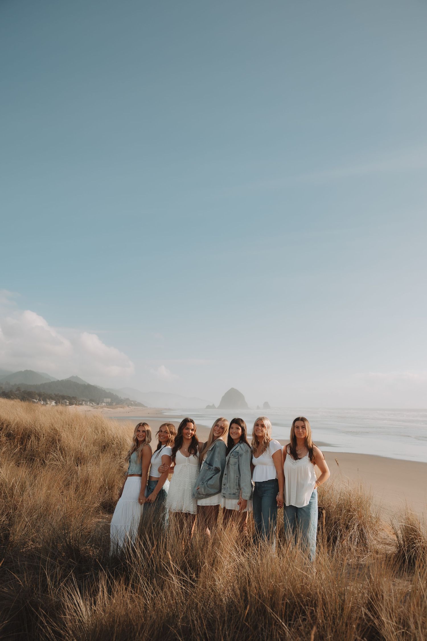Group of seven women posing together in white outfits on coastal sand dunes at Cannon Beach Oregon.