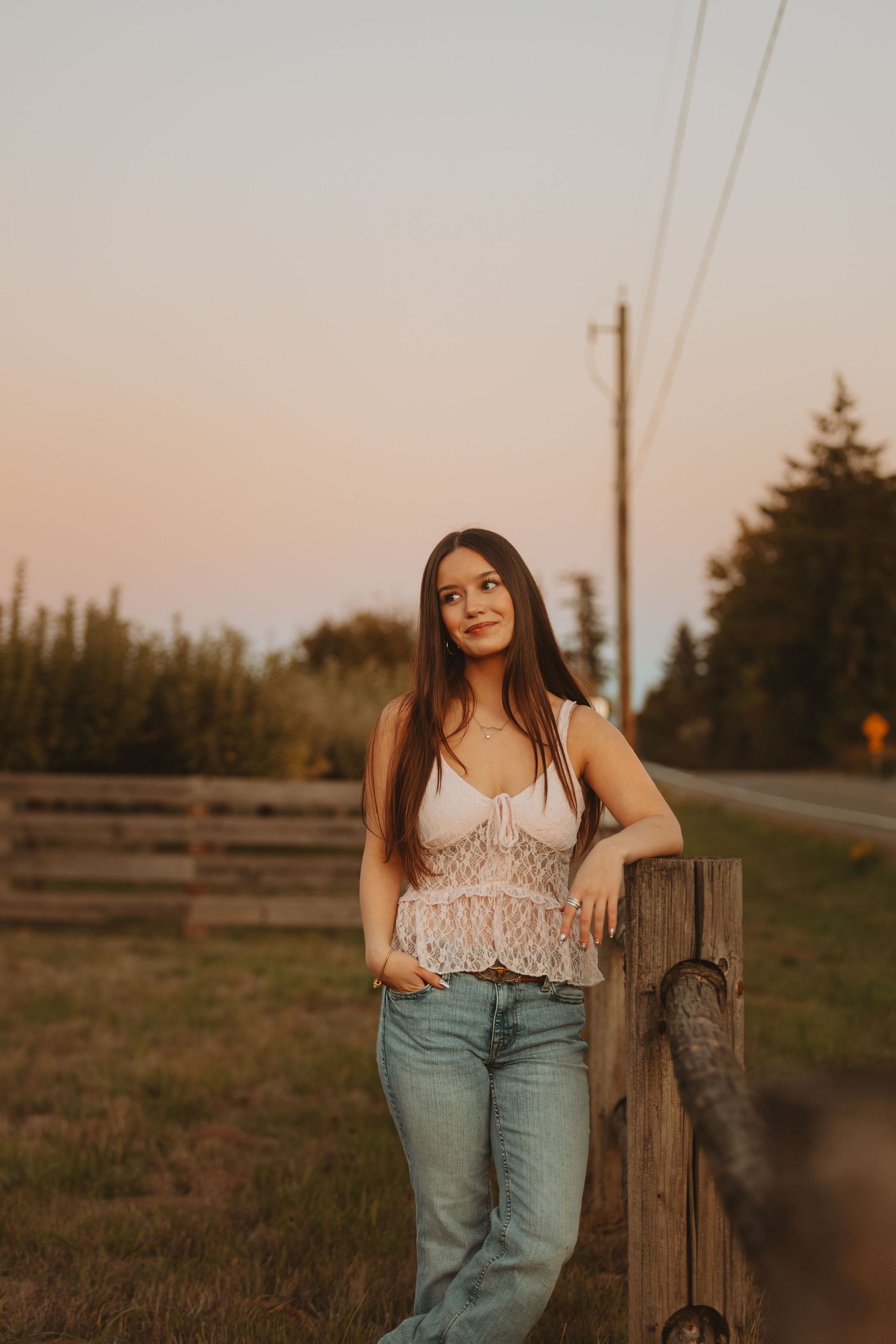 Young woman in white lace top and jeans leaning on wooden fence post at golden hour sunset in rural setting.