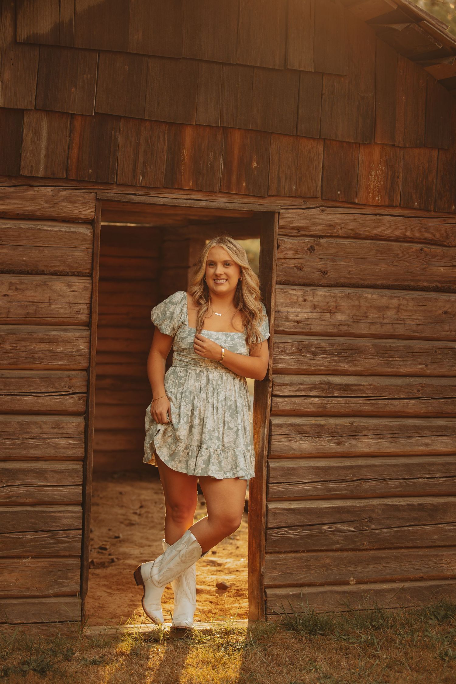Young woman in floral dress and white cowboy boots posing in rustic wooden barn doorway at golden hour.