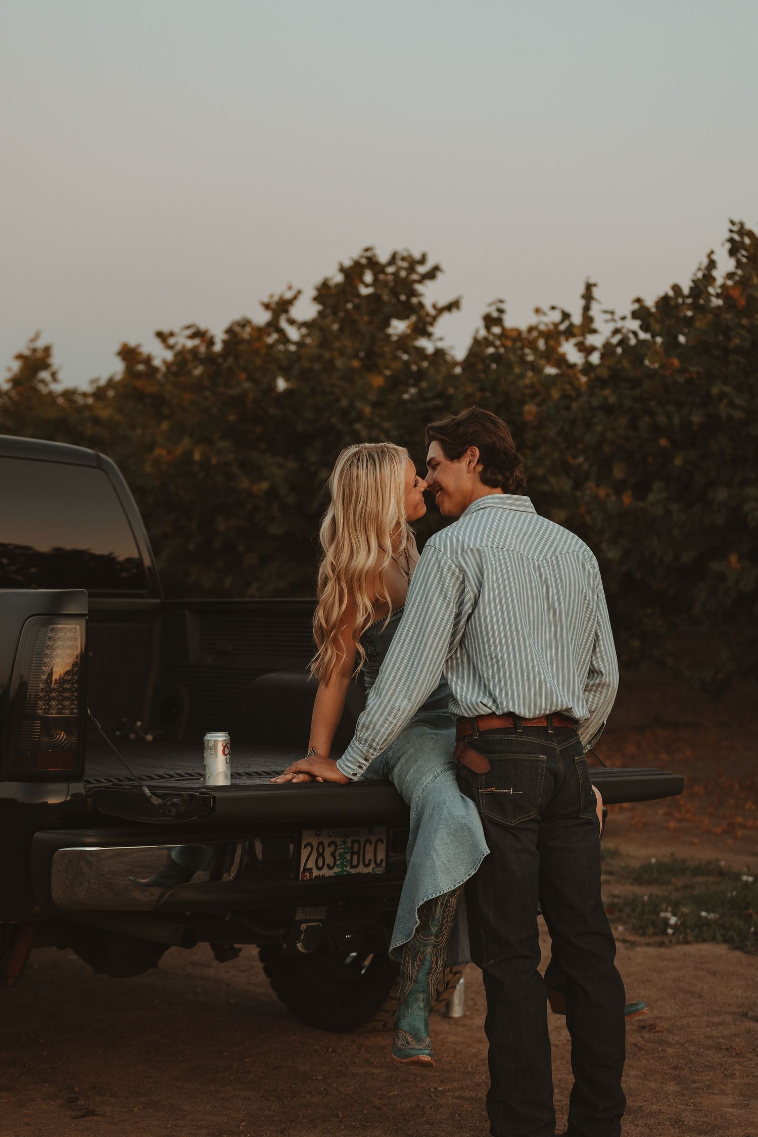 Couple leans against black pickup truck during sunset in rural setting.