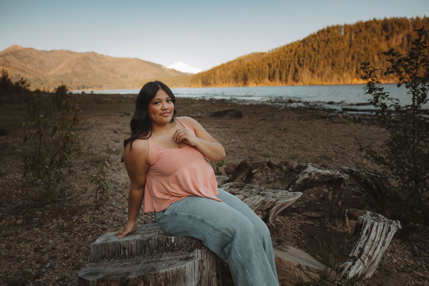 Woman in pink tank top and jeans sits on driftwood by a mountain lake at golden hour sunset.