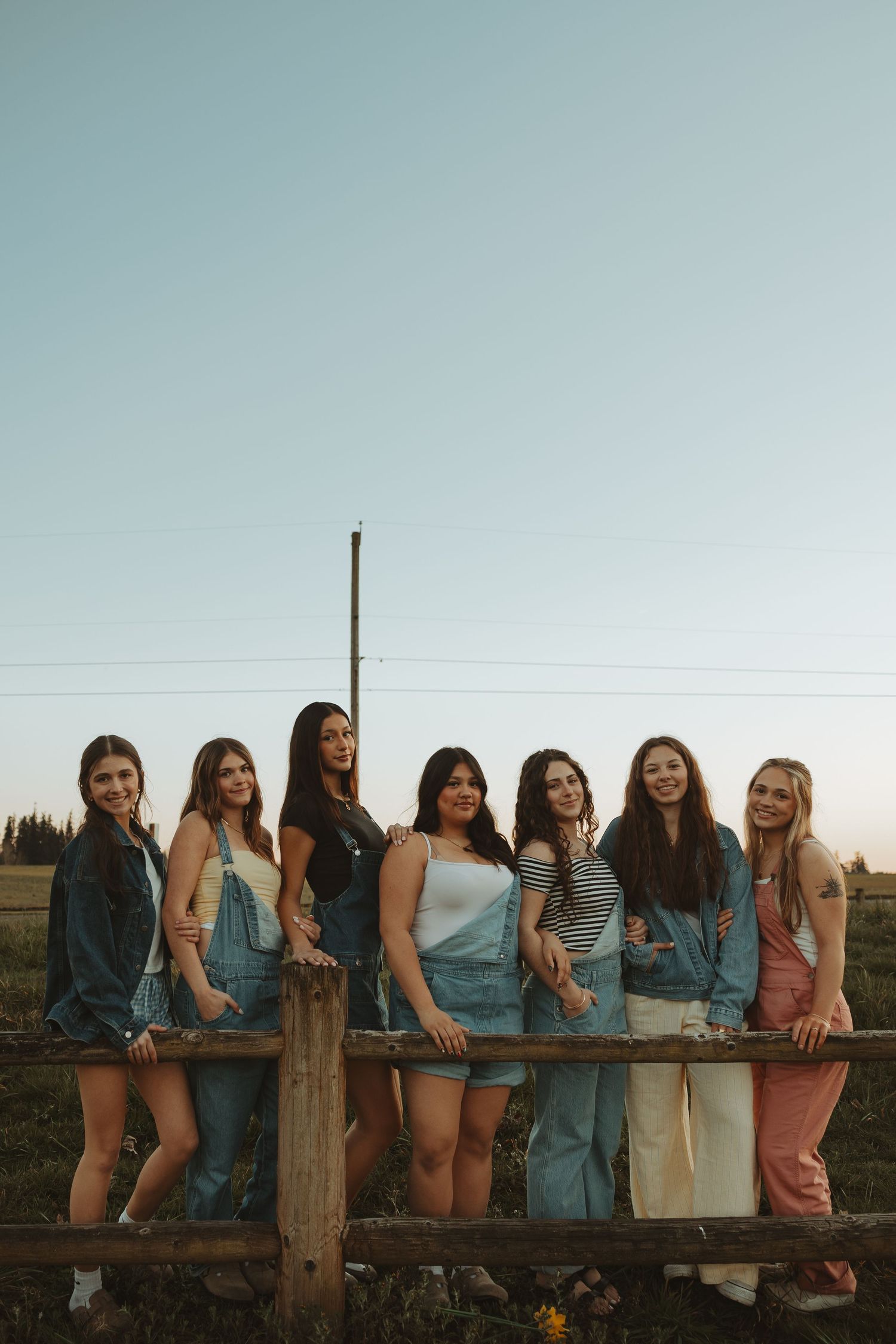 Seven young women in denim overalls pose together smiling at a rustic wooden fence during golden hour.