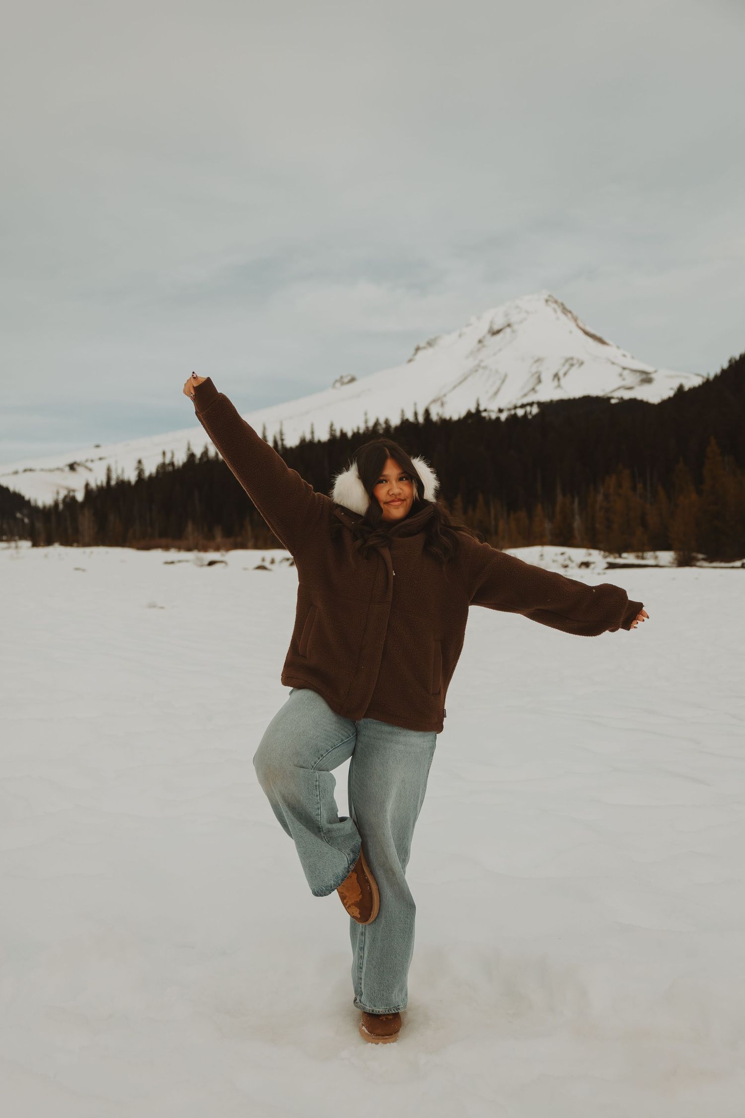 Woman in brown jacket and jeans poses playfully on one leg in a snowy mountain landscape.