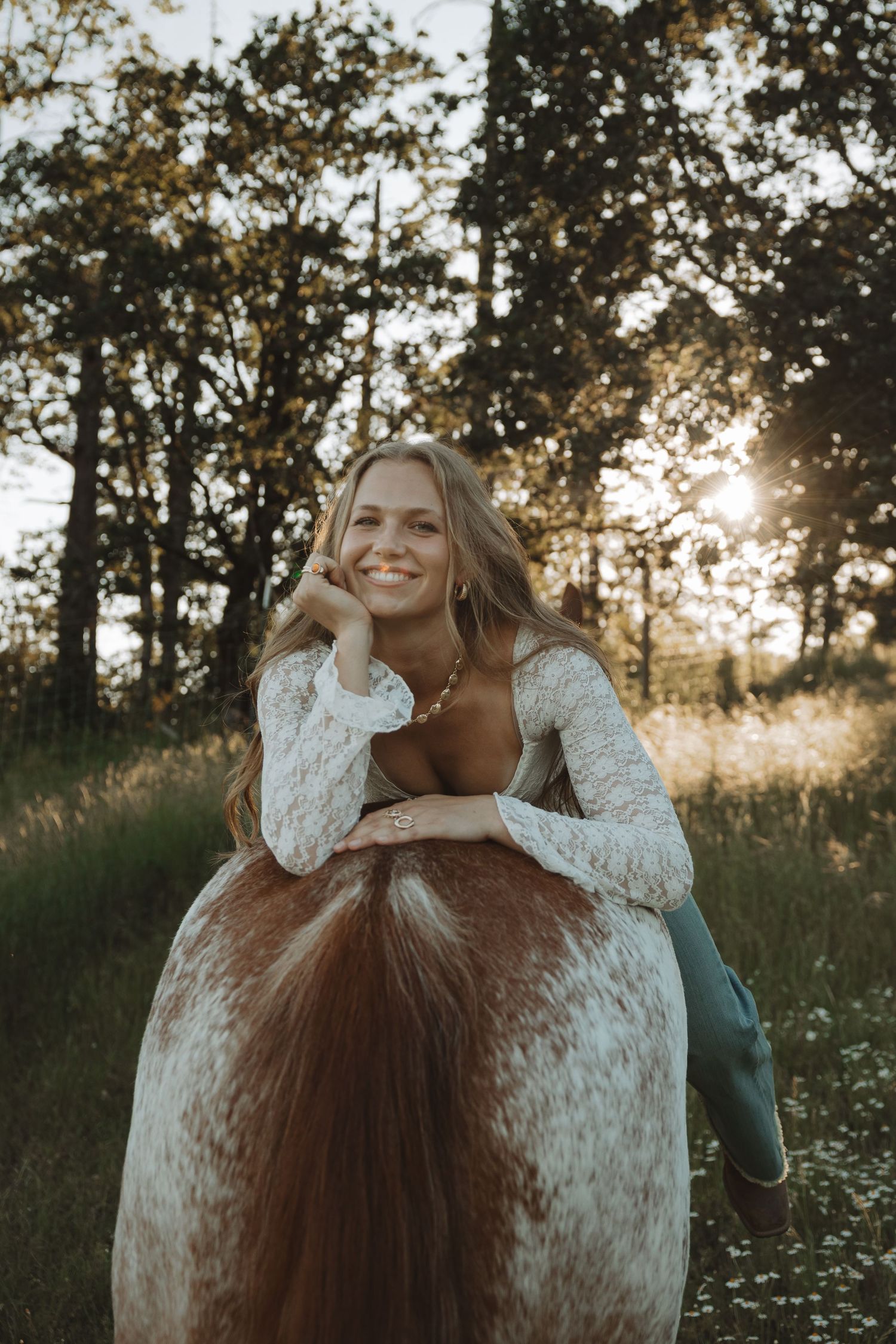 Smiling young woman leaning on a horse in a sunlit forest clearing, wearing a white lace dress at golden hour.
