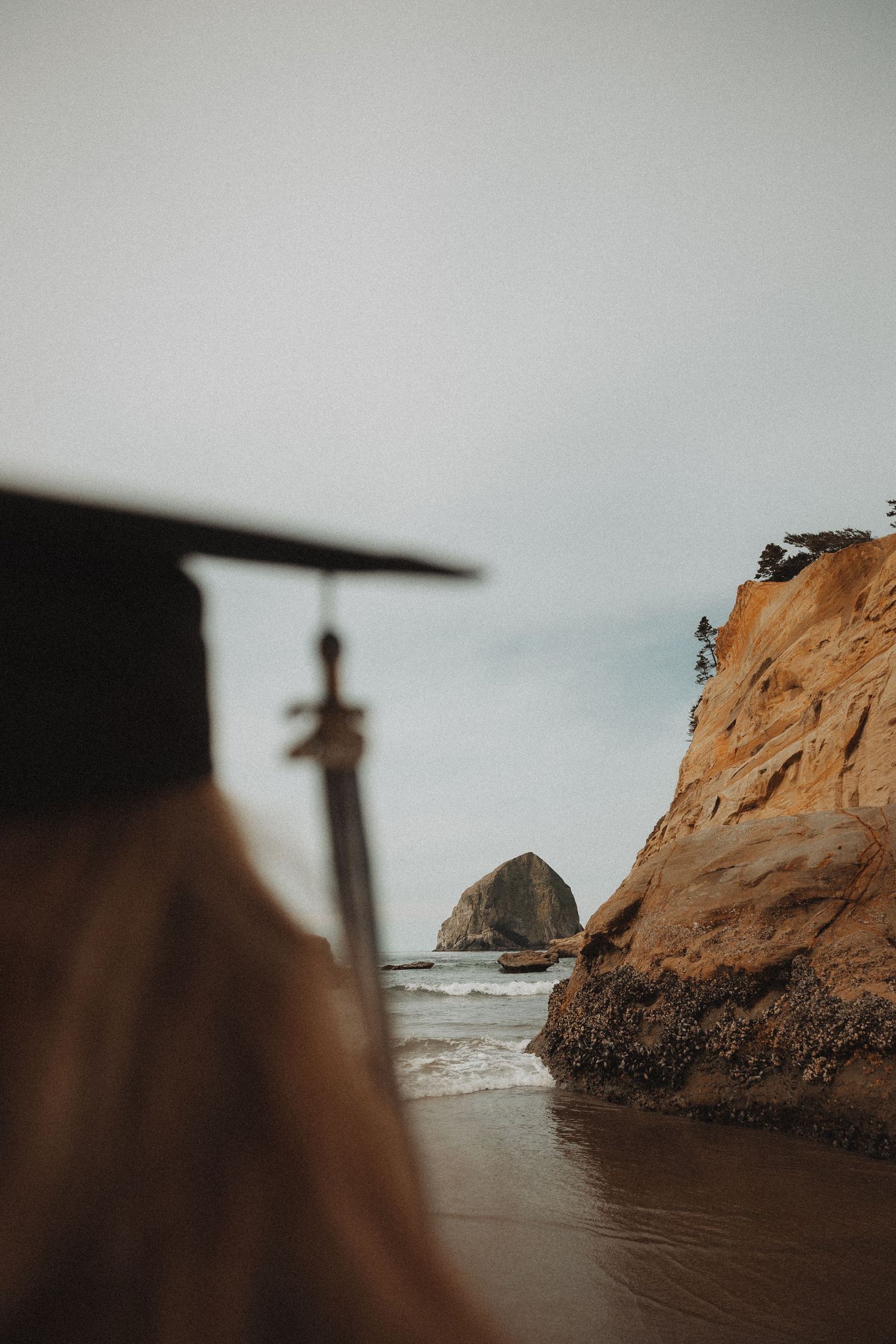 Graduate in cap stands before scenic Haystack Rock at Cannon Beach, Oregon, with dramatic cliffs and ocean waves.