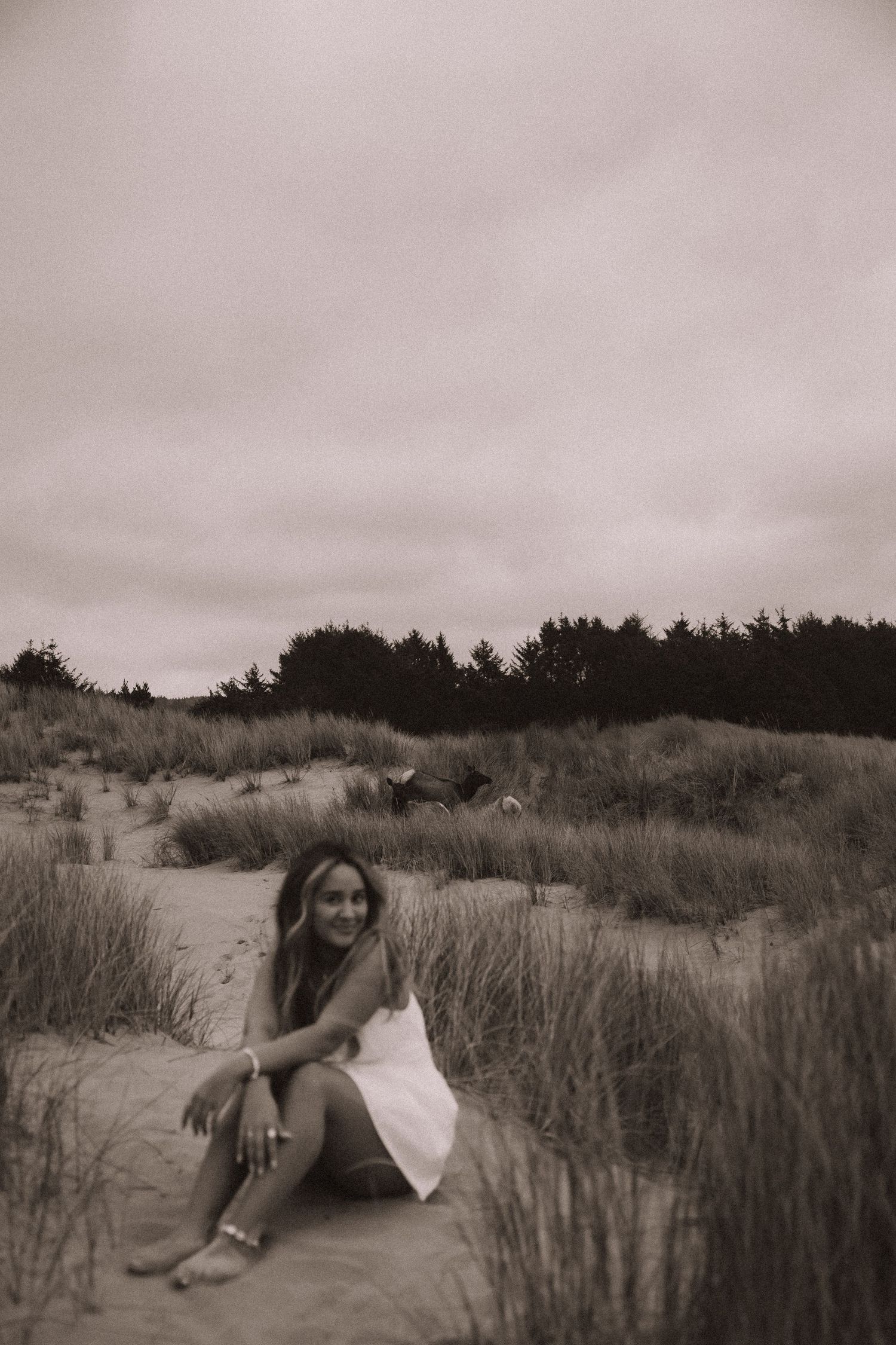 Young woman in white dress sitting on sandy dunes with coastal grass in black and white beach photo.