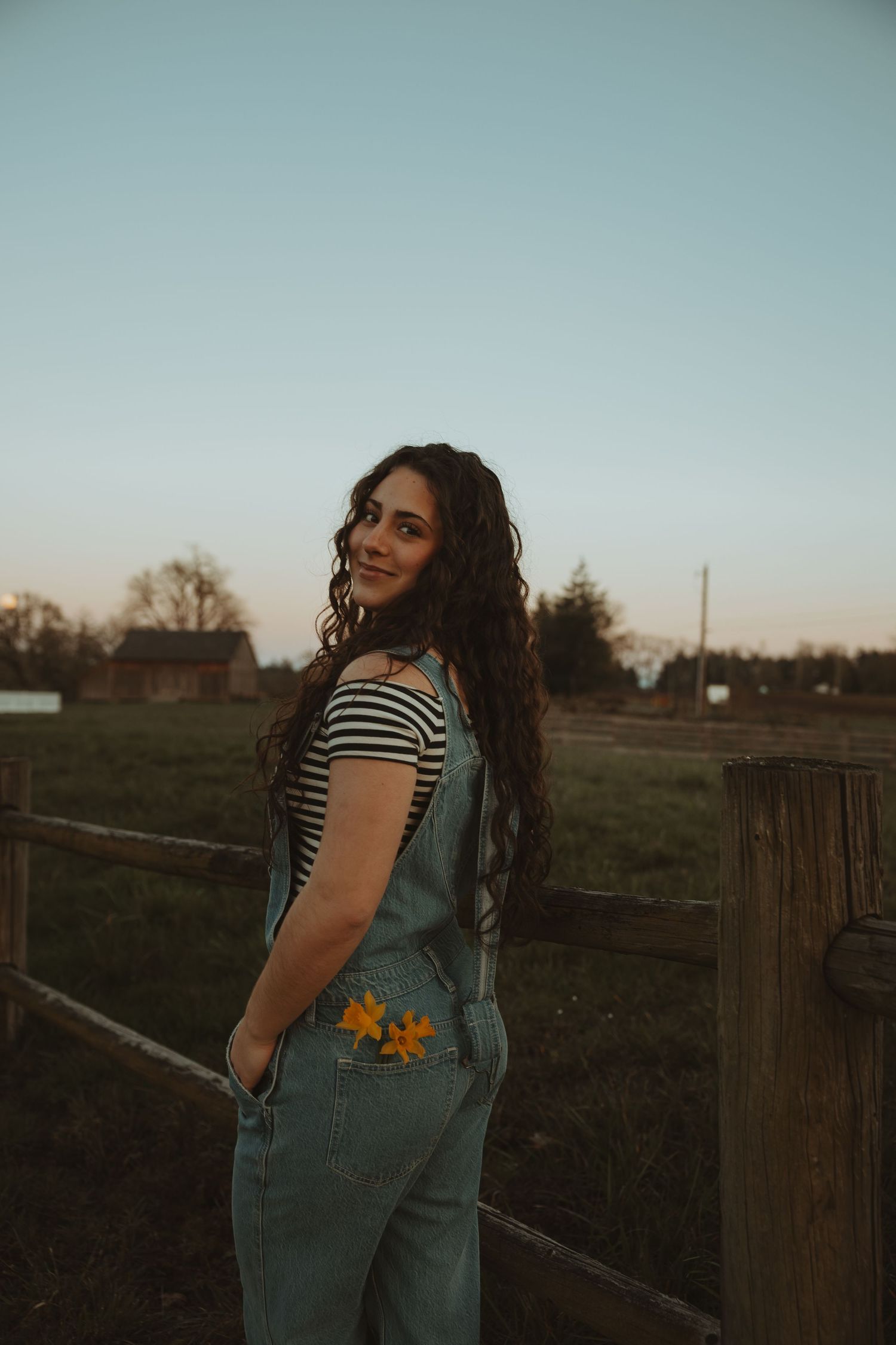 Smiling woman with curly hair in denim overalls stands by wooden fence at dusk.