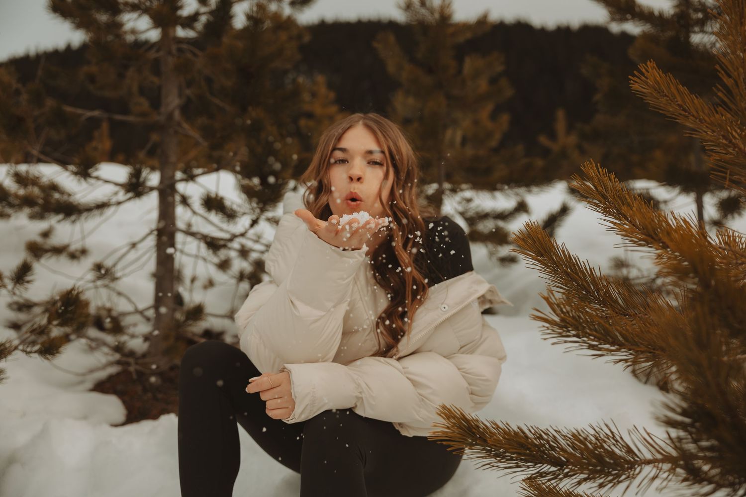 Young woman in cream puffer jacket blowing snow near pine trees in winter.