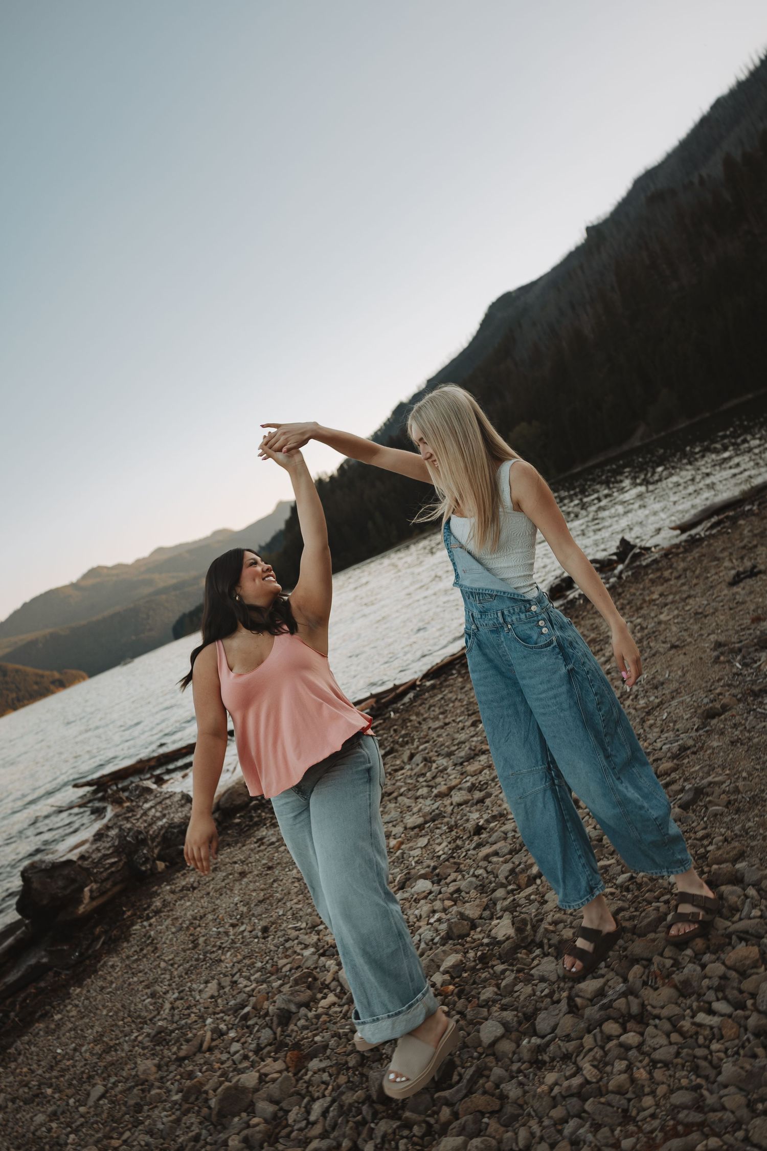 Two young women dancing together on a rocky shoreline beside a mountain lake at dusk.