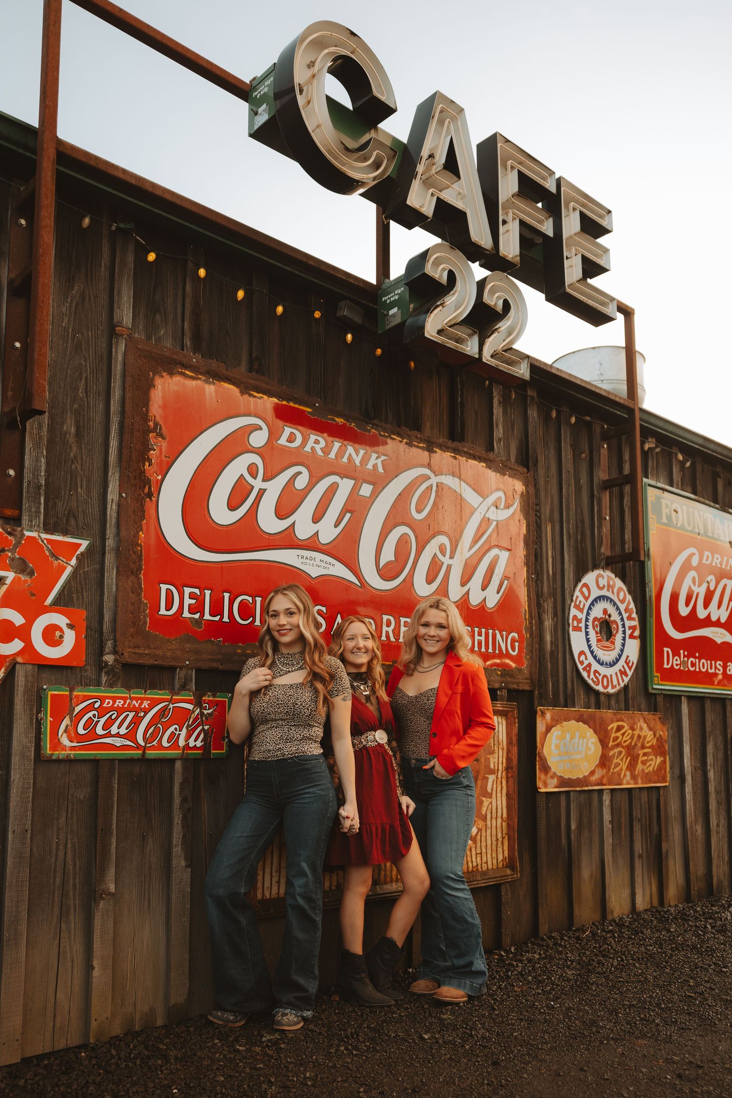 Three women pose smiling in front of Cafe 22 with vintage Coca-Cola signs on a rustic wooden wall.