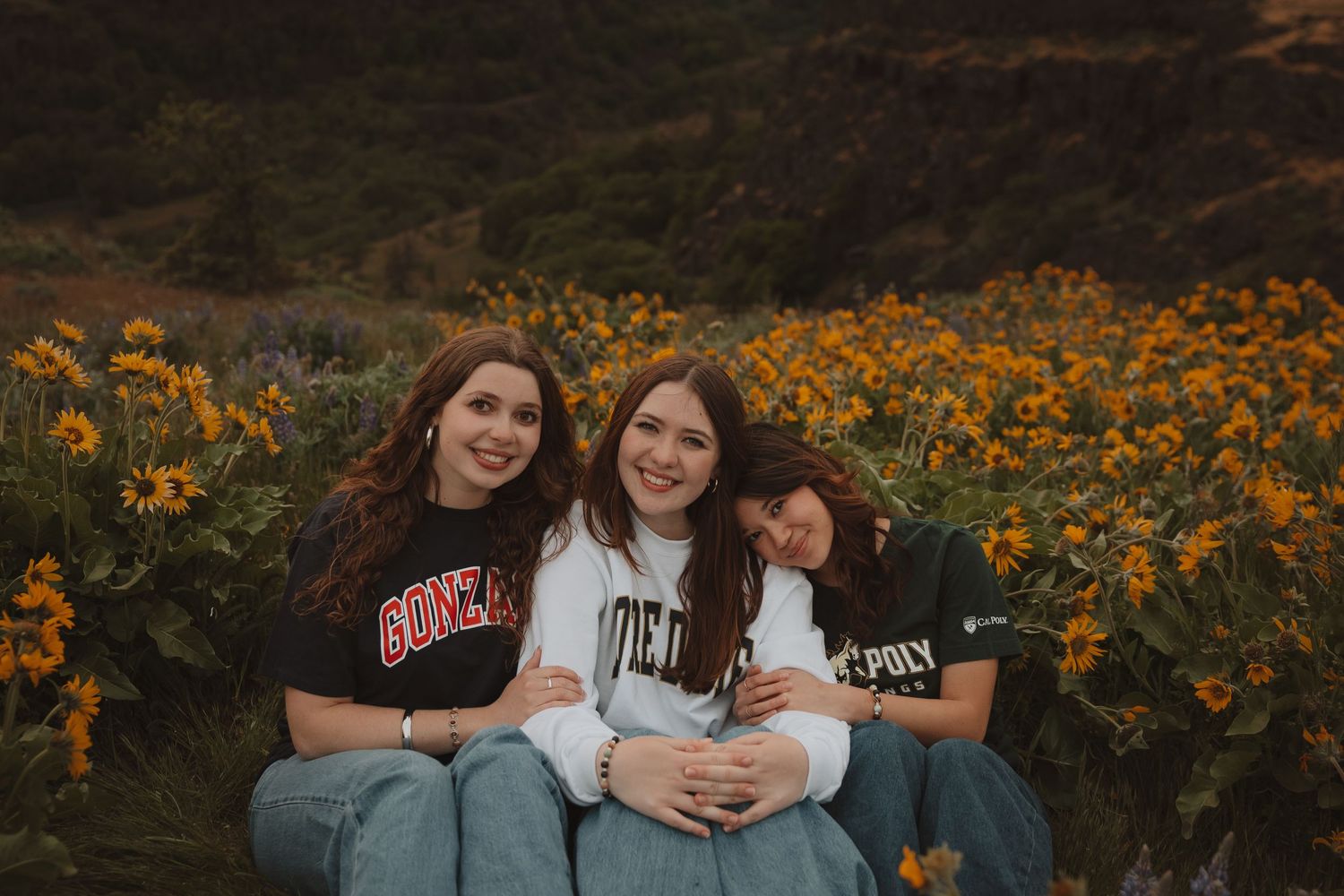 Three young women smile together in a field of yellow wildflowers, wearing college sweatshirts in a scenic outdoor setting.
