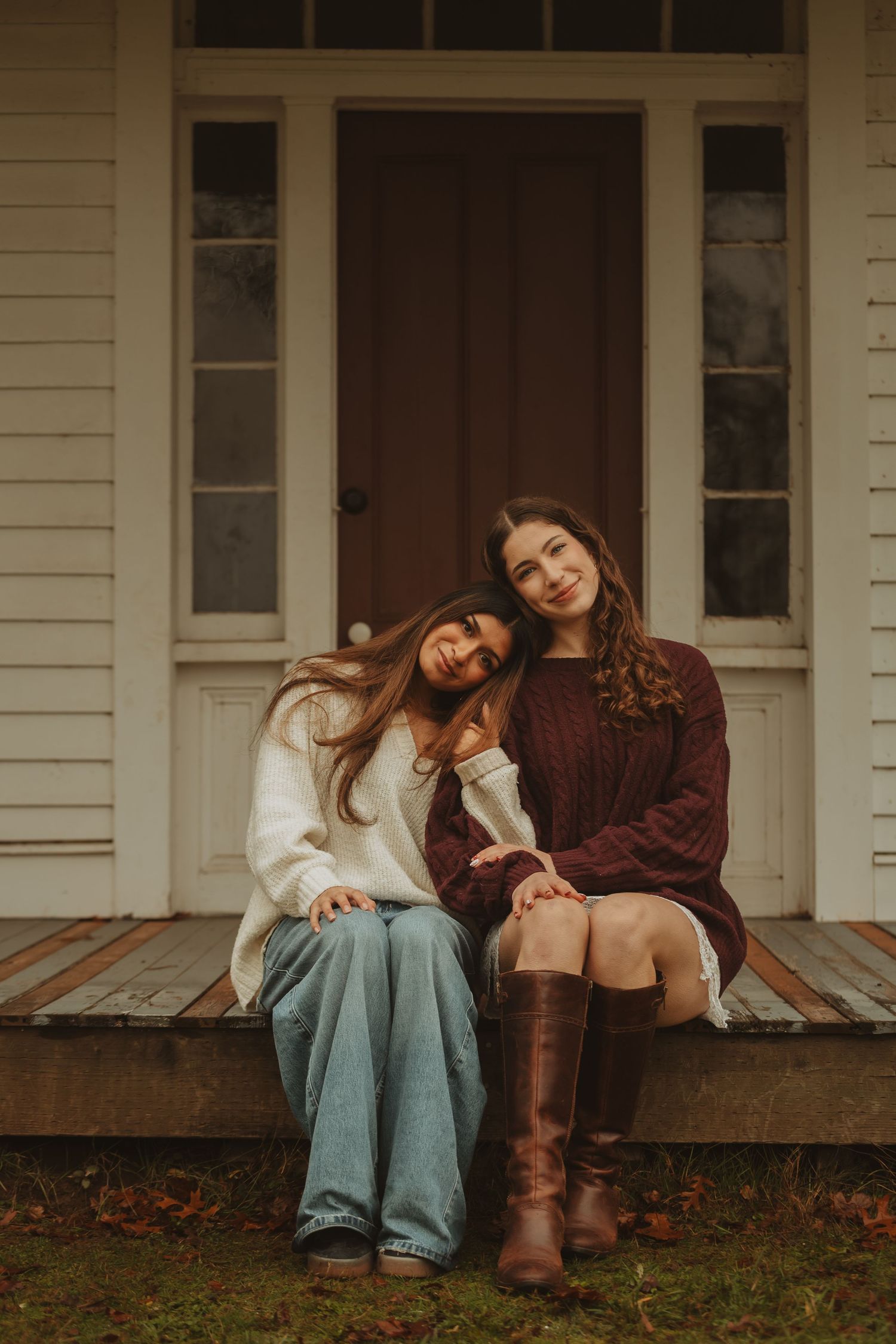 Two people in casual attire relax on wooden steps of a white farmhouse porch.